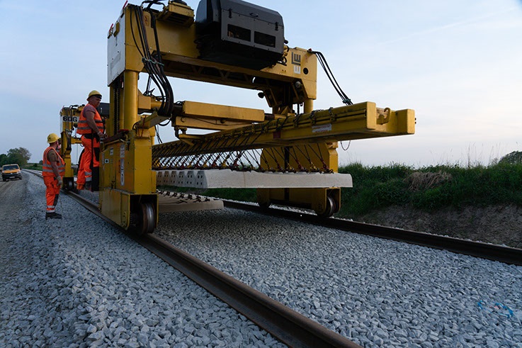 Eine Gleisbau-Maschine arbeitet an einer Schotterbahn, während mehrere Arbeiter in Sicherheitskleidung in der Nähe stehen.