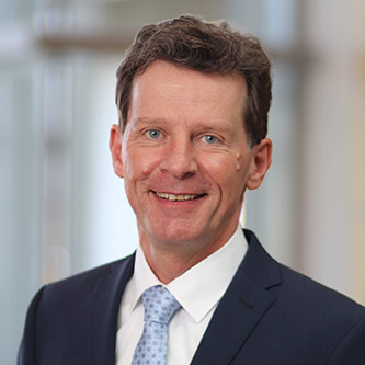 Ein lächelnder Mann in einem Anzug mit einer hellblauen Krawatte steht vor einem unscharfen Hintergrund.