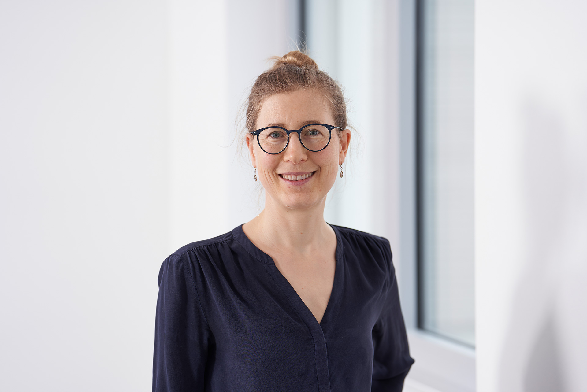 Eva Hartmann, lächelnd mit Brille vor einem hellen Hintergrund und einem Fenster stehend.