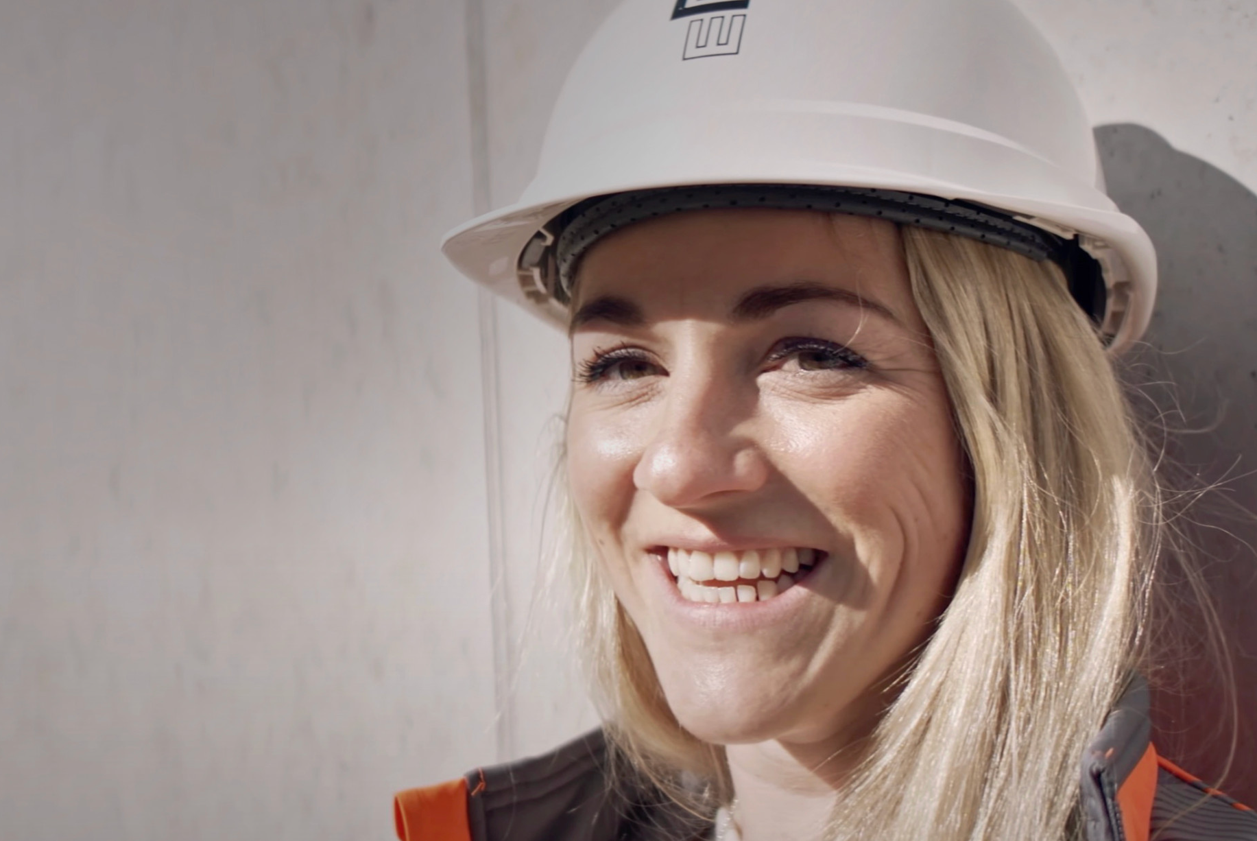 Eine blonde Frau in einem weißen Helm und einer orangefarbenen Jacke lächelt fröhlich, während sie an einer hellen Wand steht.