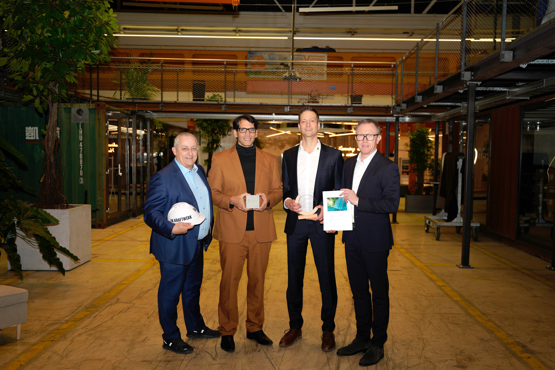 Drei Männer in Anzügen halten Papiere und stehen in einem Industriegebäude.