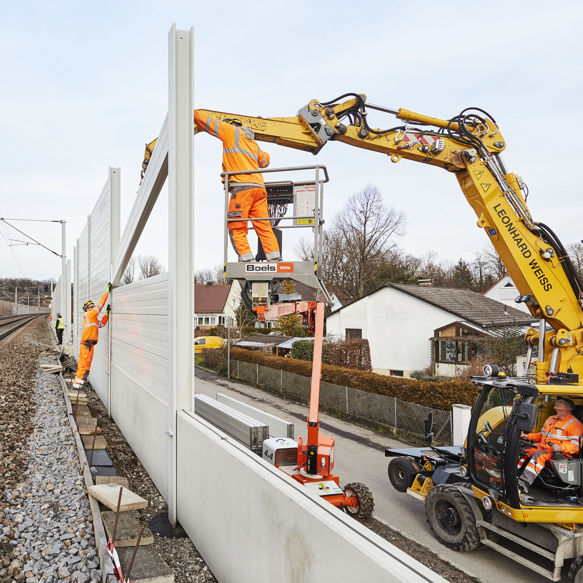 Arbeiter in leuchtend orangefarbener Kleidung montieren eine Lärmschutzwand entlang einer Bahnstrecke, während ein Kran hilft.