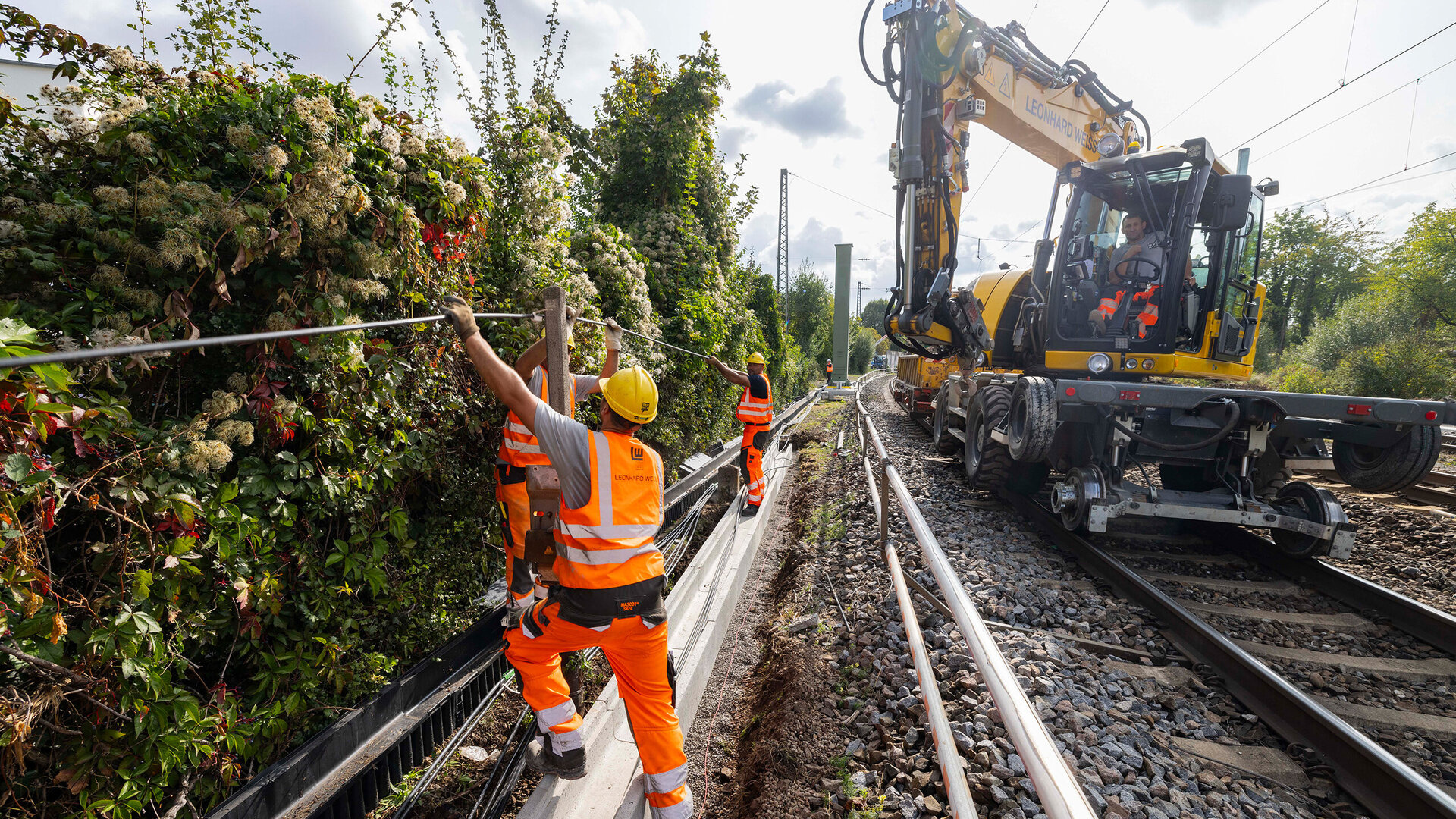 Mitglieder eines Teams in Sicherheitskleidung arbeiten an der Beseitigung von Gestrüpp neben den Gleisen, während ein Kranfahrzeug bereitsteht.