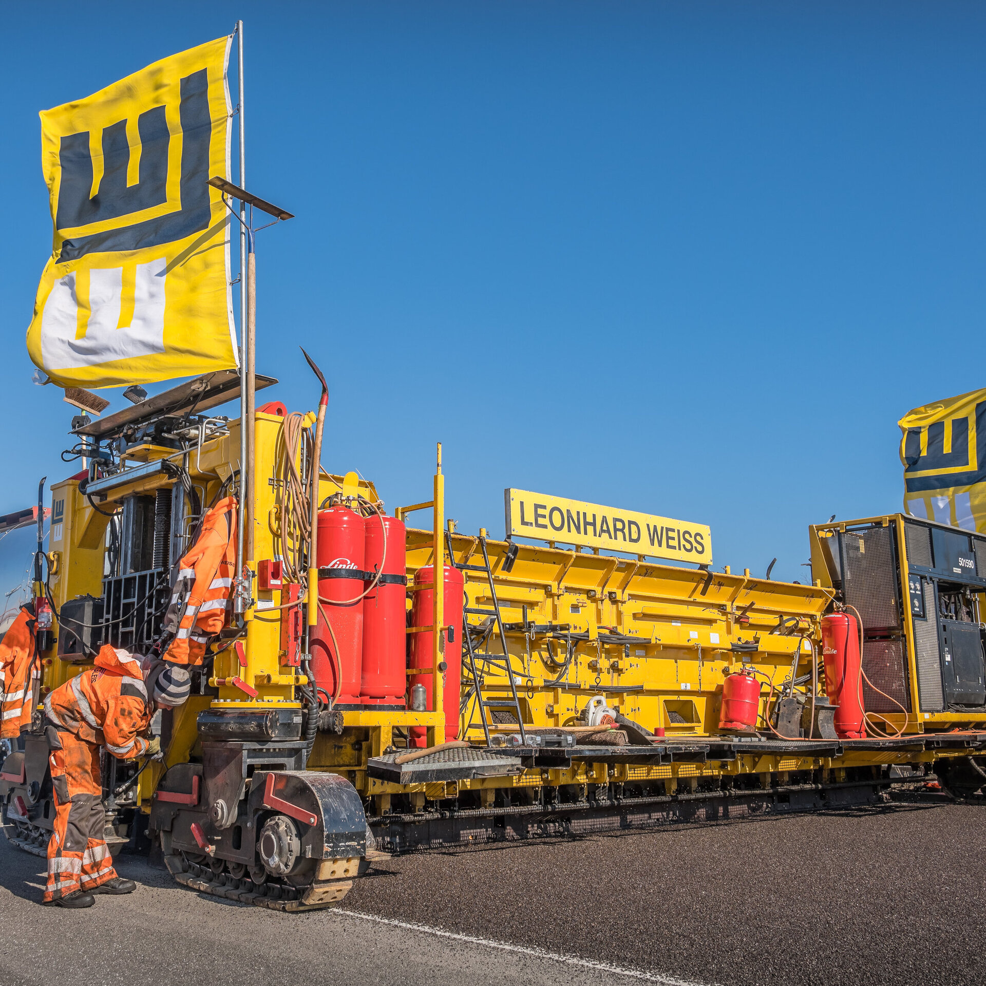 Ein Straßenbaufahrzeug mit dem Namen Leonhard Weiss wird von mehreren Arbeitern in orangefarbener Kleidung inspiziert.