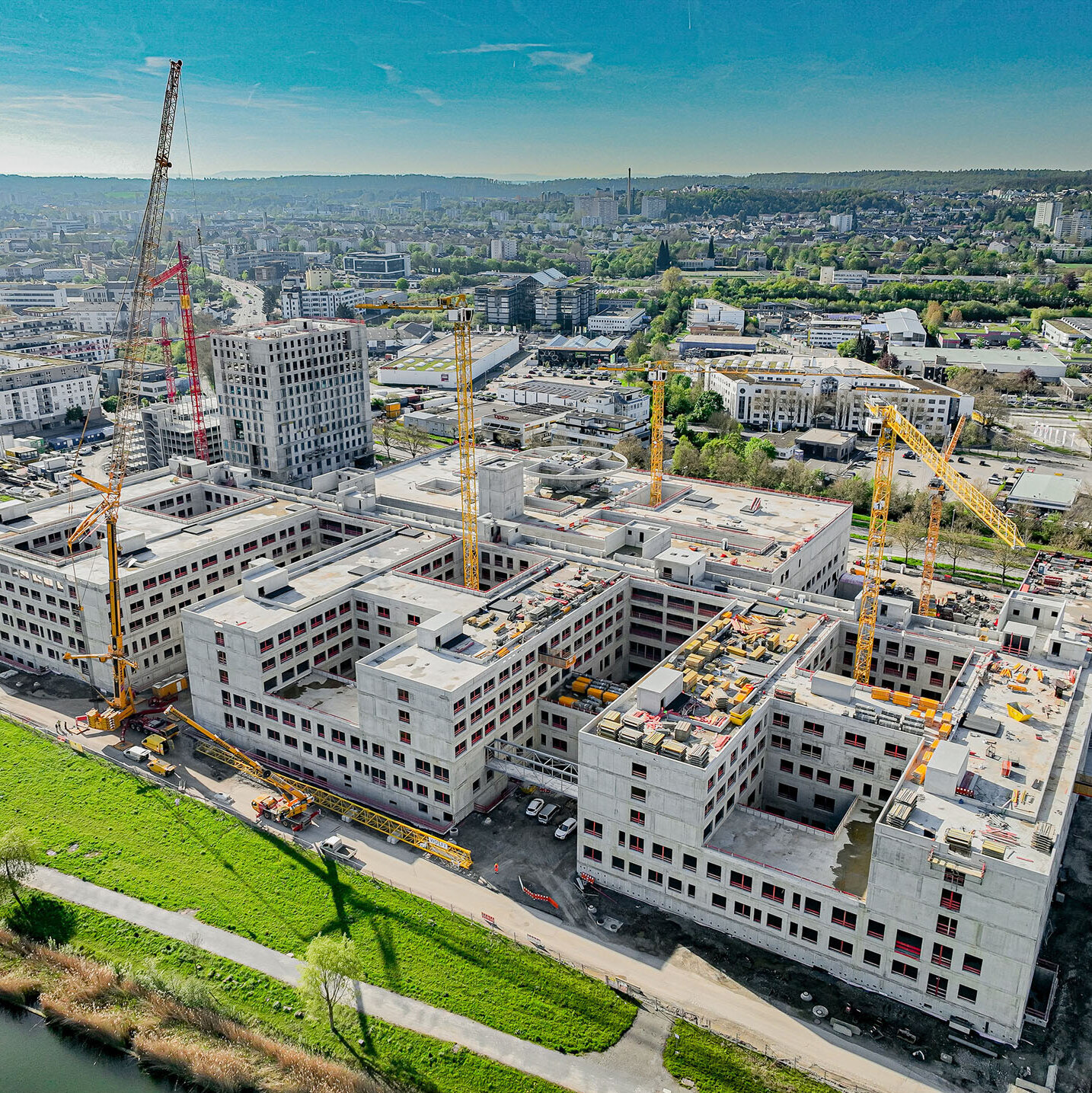 Kranarbeiten an einem großen Bauprojekt, während die Stadt im Hintergrund sichtbar ist, unter einem klaren blauen Himmel.