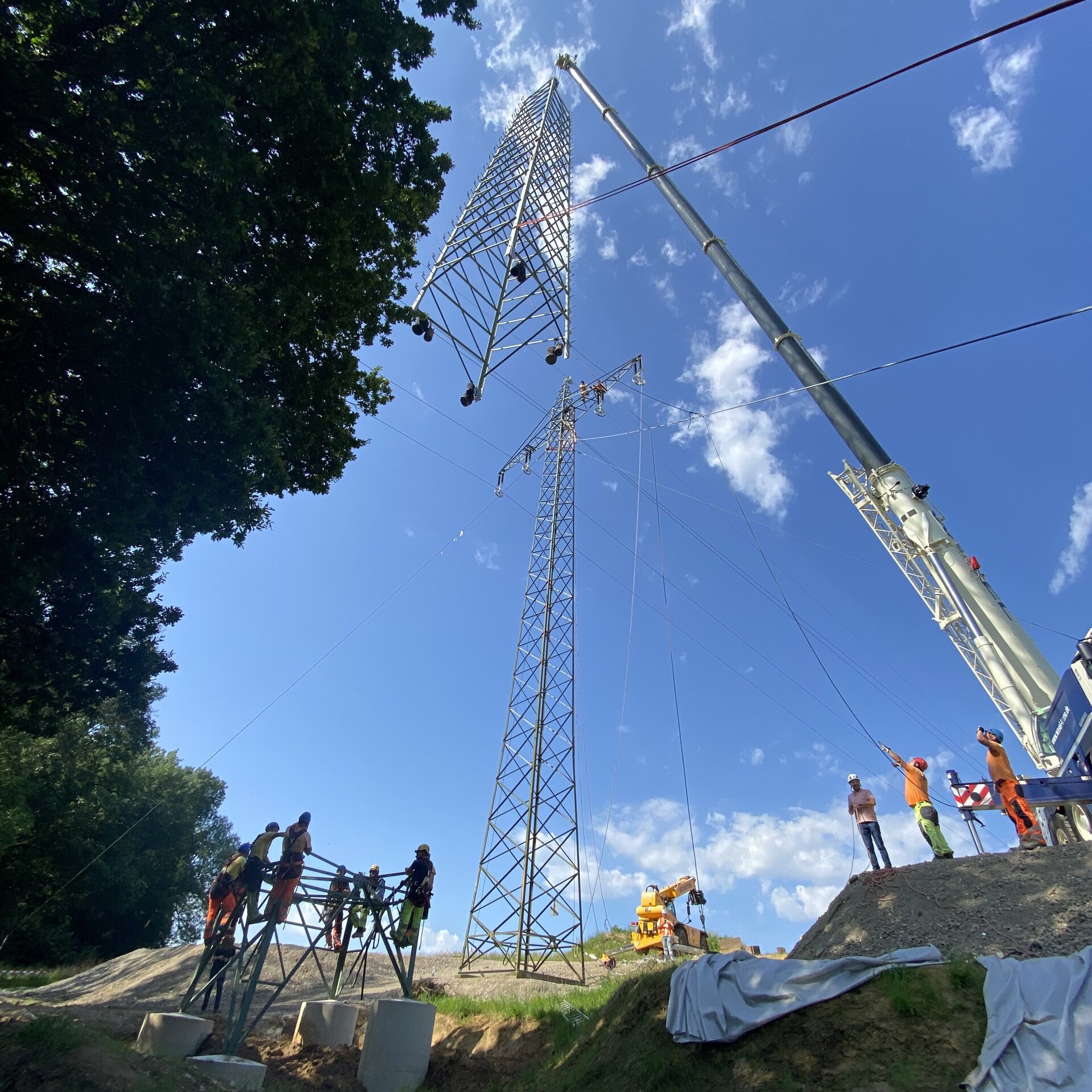 Ein Kran hebt eine Struktur in die Höhe, während mehrere Arbeiter in orangefarbener Kleidung an einem Strommast arbeiten.
