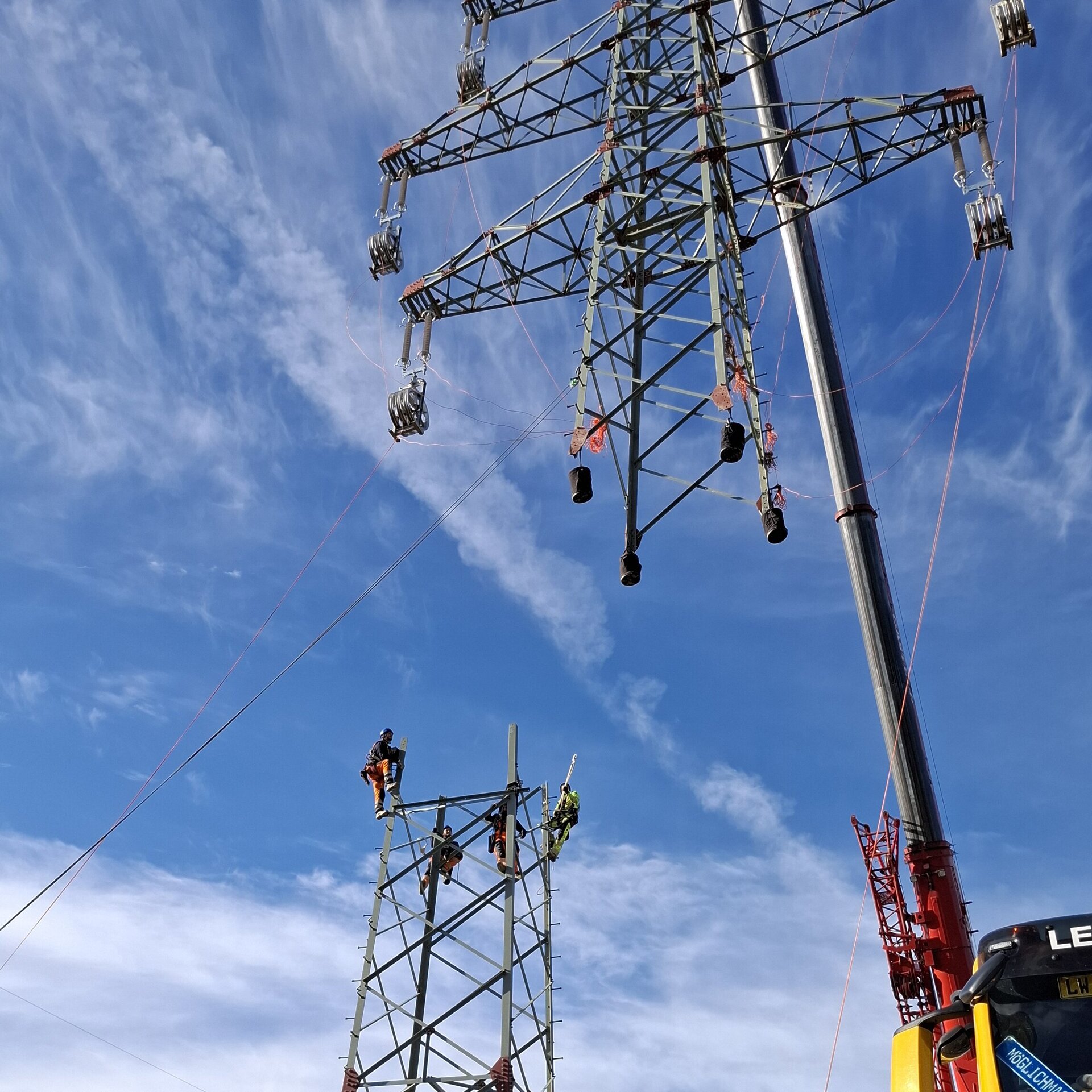Ein Mitarbeiter steuert einen Kran, der die Spitze eines Strommastes anhebt, während Arbeiter am unteren Teil bereits warten, um die Spitze anschließend zu montieren.