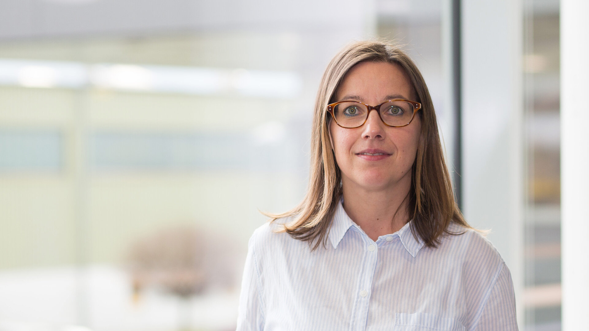Eine Frau mit langen, braunen Haaren steht an einem Geländer und lächelt, während sie in die Kamera schaut.