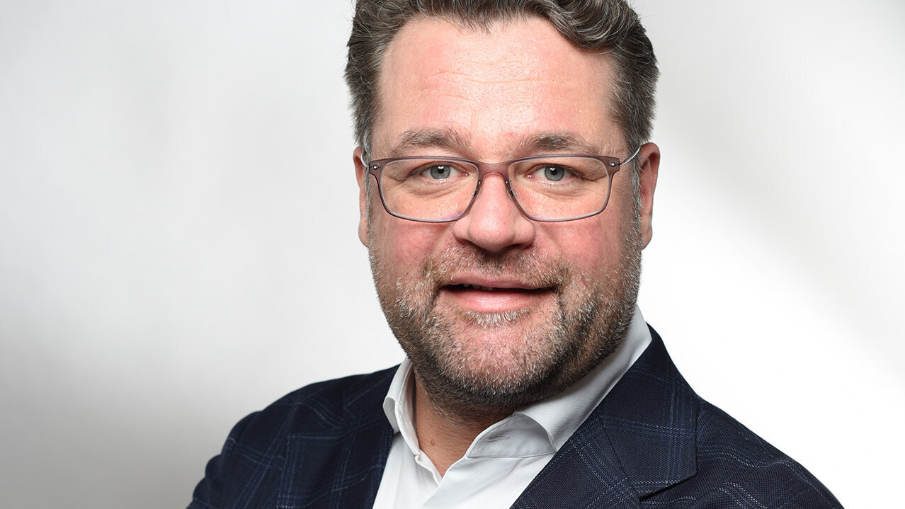 Ein Mann mit Brille und Bart lächelt freundlich, während er seine Arme vor der Brust verschränkt.