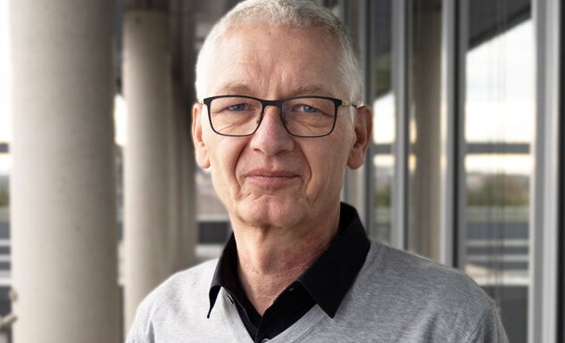 Ein selbstbewusster älterer Herr in einem grauen Pullover steht an einer modernen Gebäudefront mit einem minimalistischen Stil.