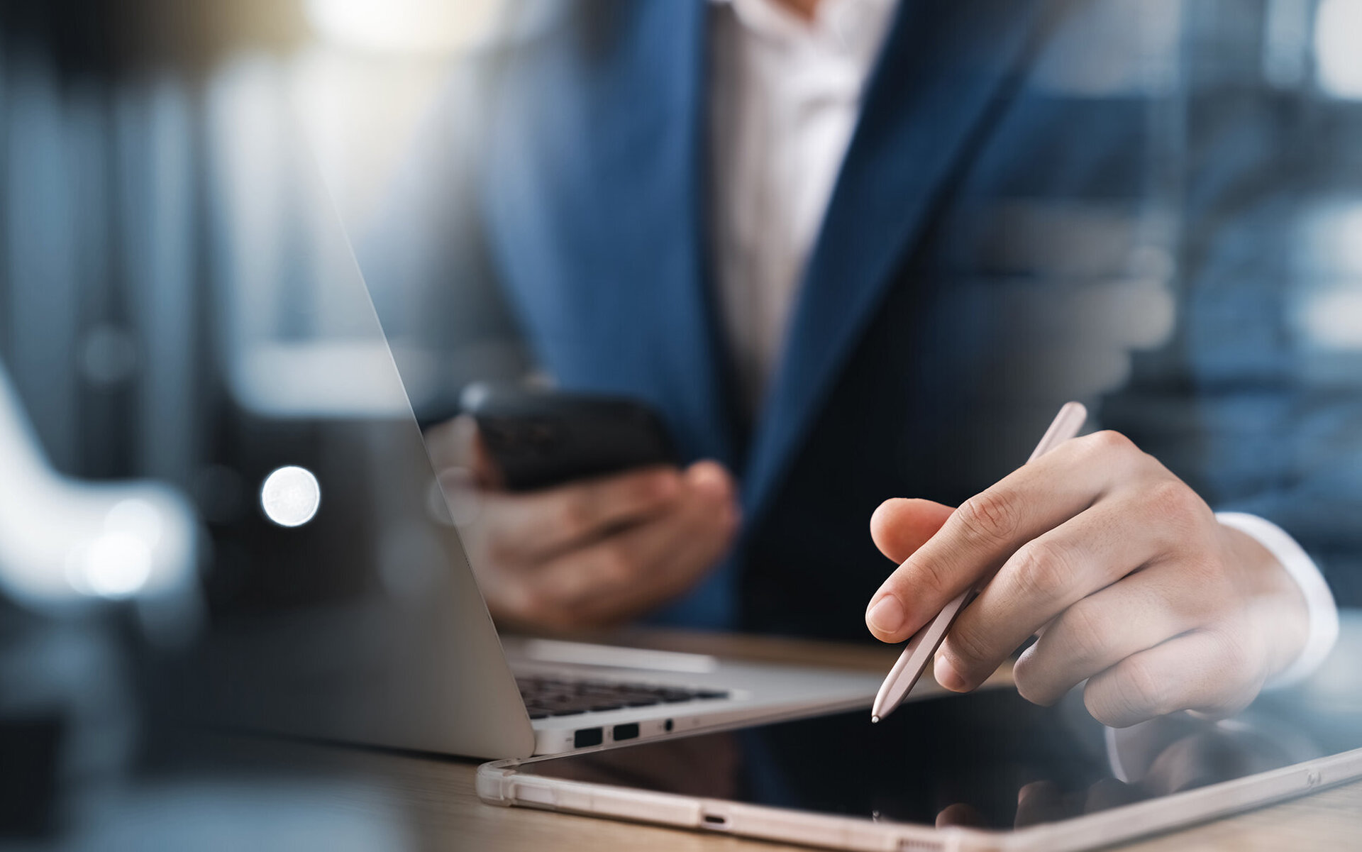 Der Fokus liegt auf der Hand eines Mannes, die einen Stift über ein Tablet führt, während ein Laptop im Hintergrund sichtbar ist.