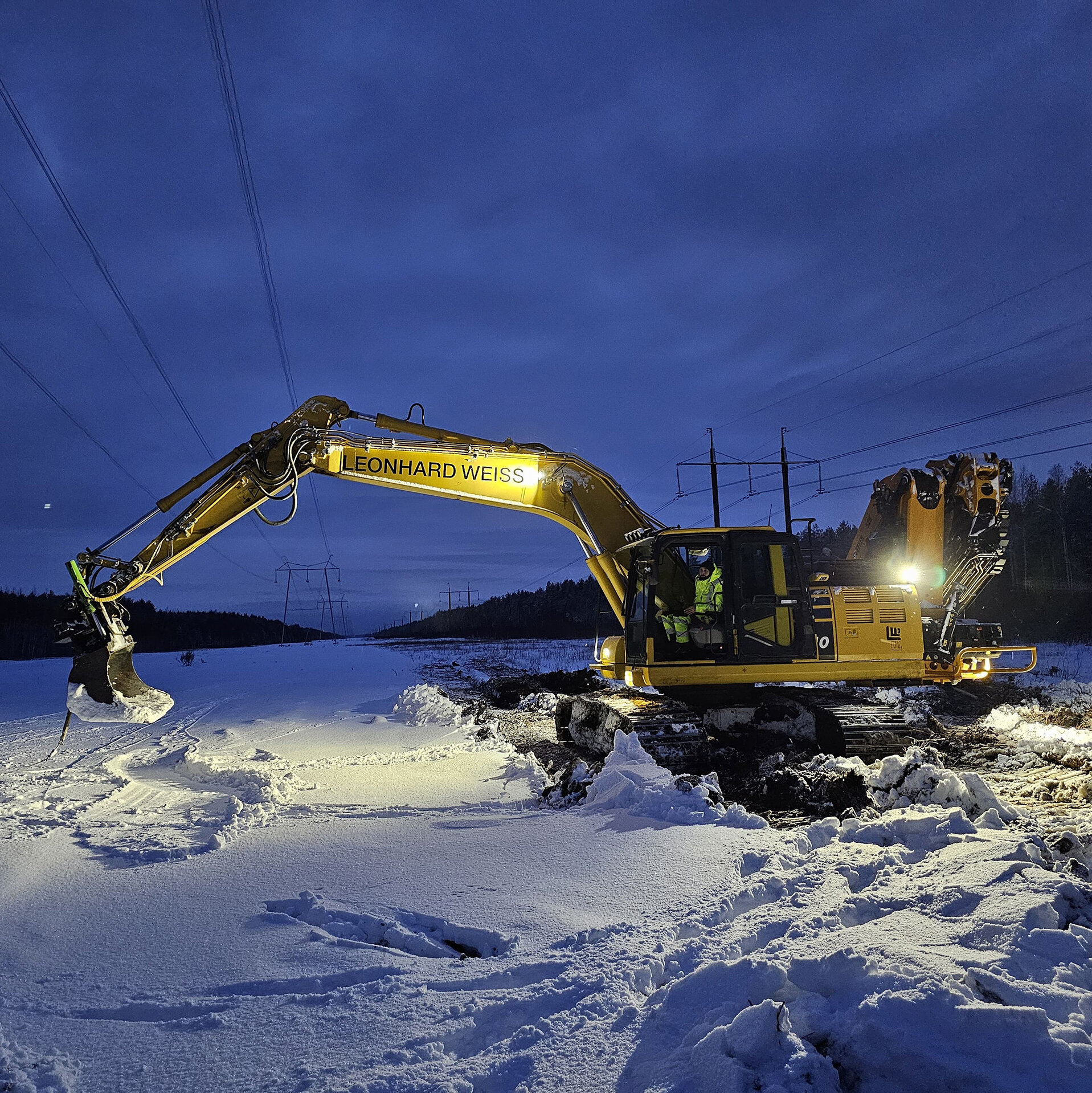 Der Bagger entfernt Schnee in der Dämmerung, während die Lichter des Fahrzeugs die Umgebung erhellen und die Arbeit erleichtern.