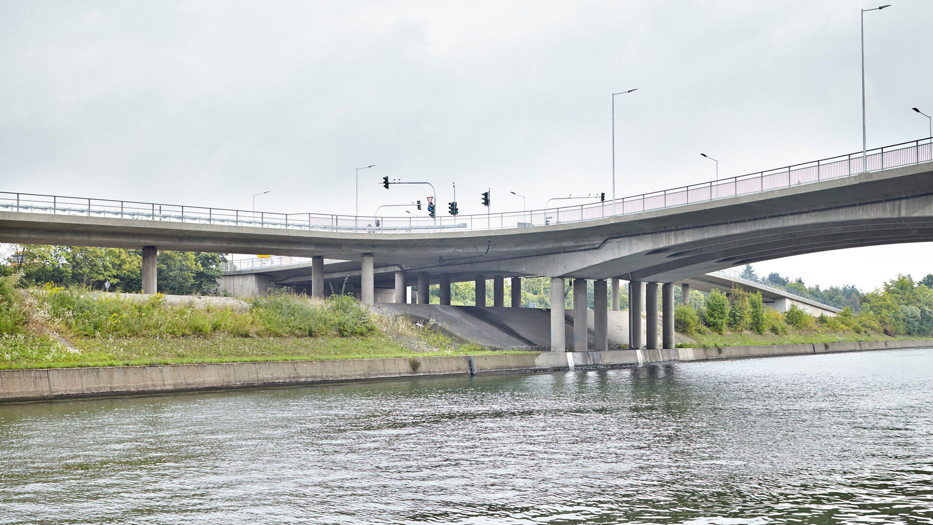 A view of a bridge crossing a river, with traffic signals and an overgrown shoreline in the background.