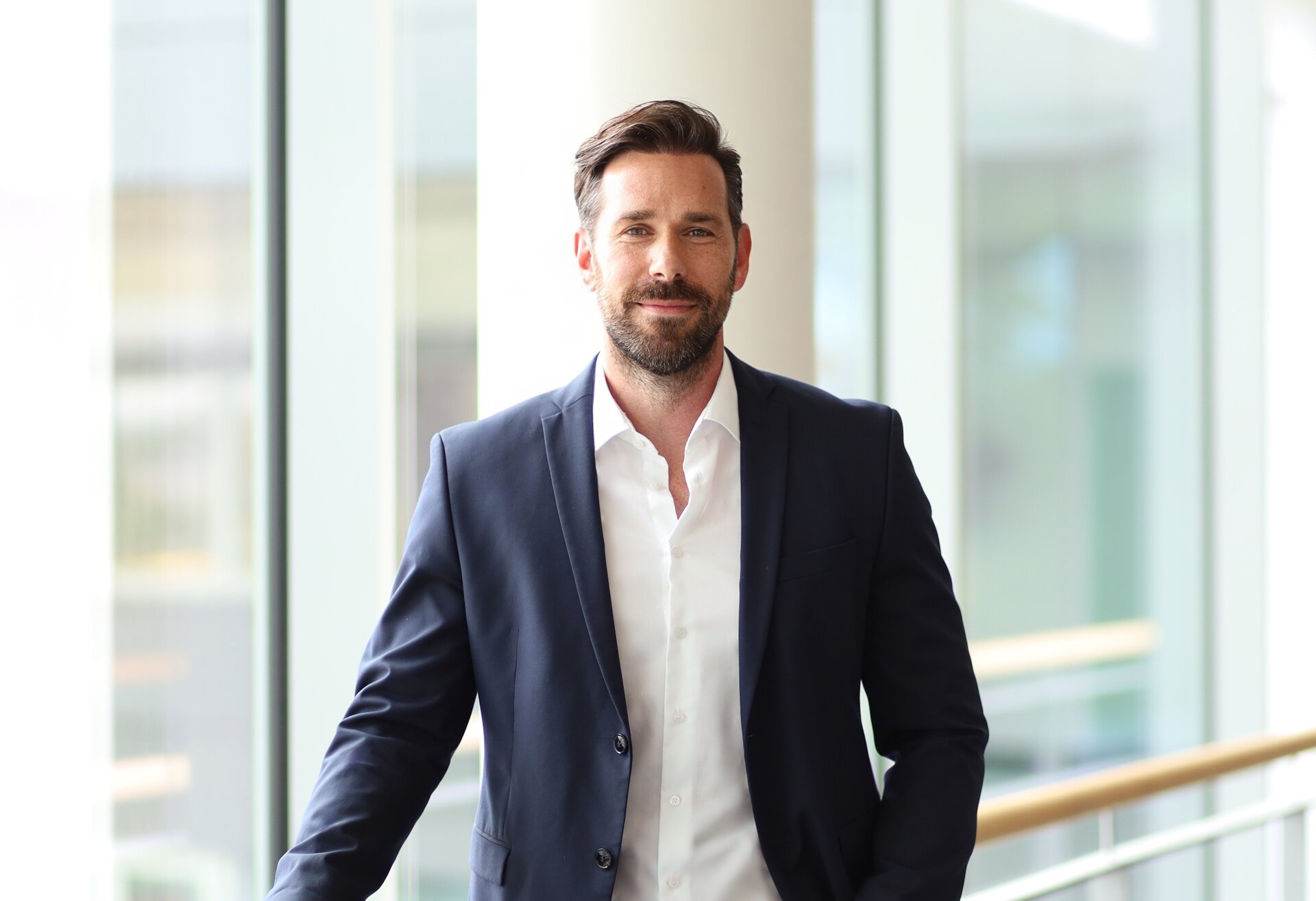 In einem stilvollen Bürogebäude posiert ein Mann in formeller Kleidung vor großen Fenstern mit natürlichem Licht.