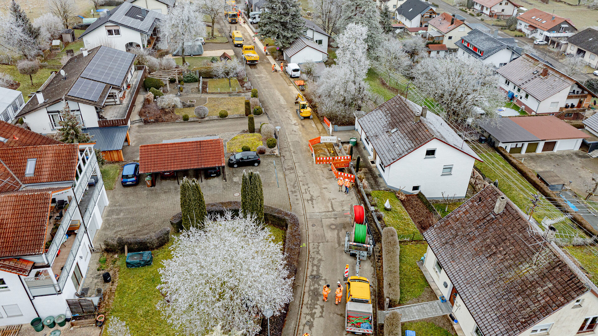 Blick auf eine ruhige Wohnstraße, wo Bauarbeiter mit orangefarbener Sicherheitskleidung an der Straße arbeiten, umgeben von frostigen Bäumen.