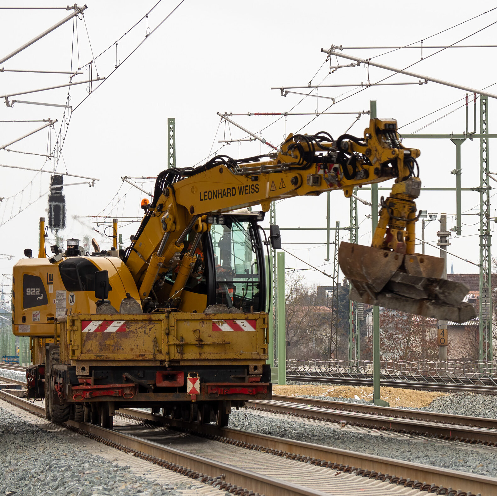 Ein gelber Bagger arbeitet an den Gleisen, während Stromleitungen über die Schienen gespannt sind und eine graue Landschaft im Hintergrund sichtbar ist.