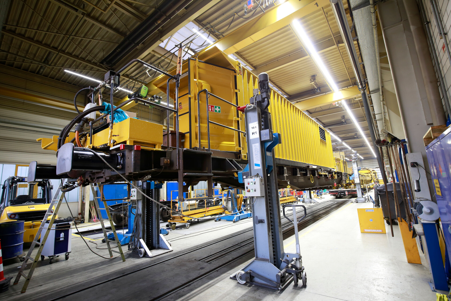 Ein gelber Zug steht in einer Gleisbauhalle, während Arbeiter an seiner Instandhaltung tätig sind.