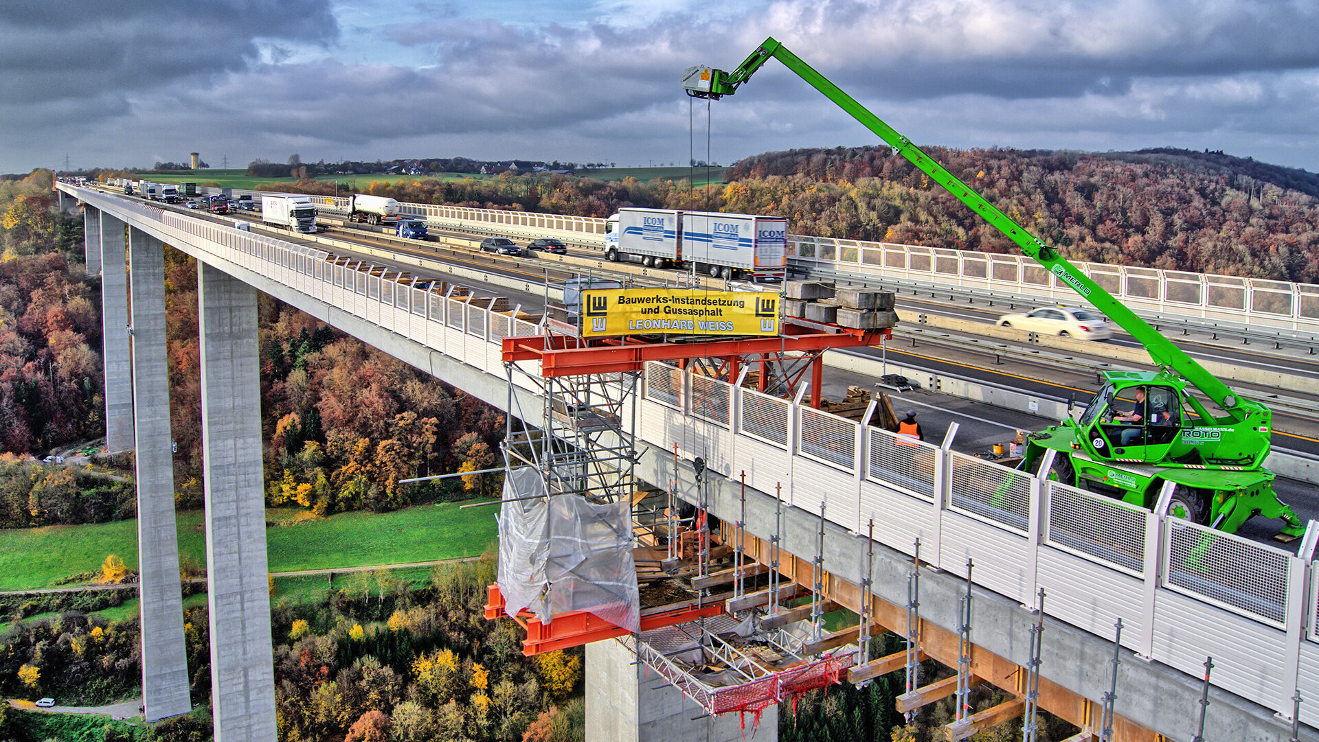 The scene shows a motorway bridge undergoing construction work, where a green crane is lifting materials and the autumnal landscape frames the scene.