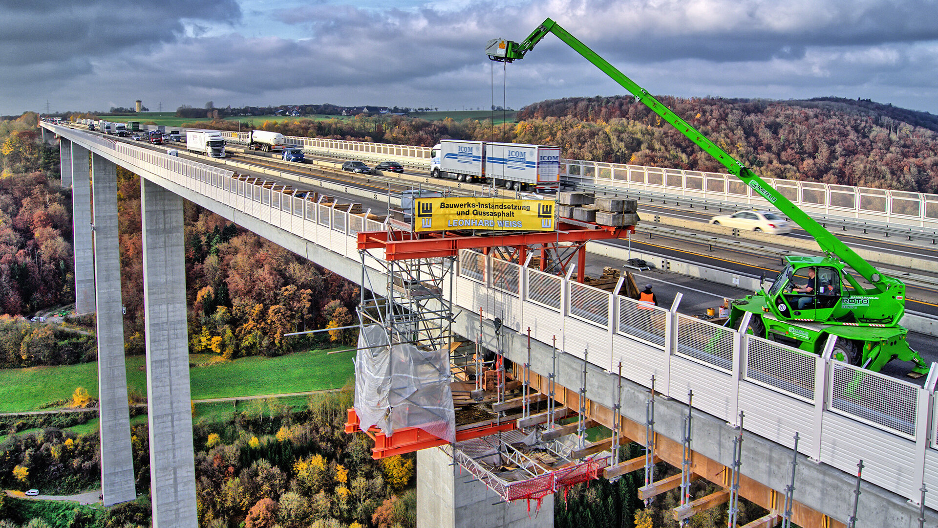 Die Szene zeigt eine Autobahnbrücke mit Bauarbeiten, wo ein grüner Kran Materialien anhebt und die herbstliche Landschaft umrahmt die Szene.