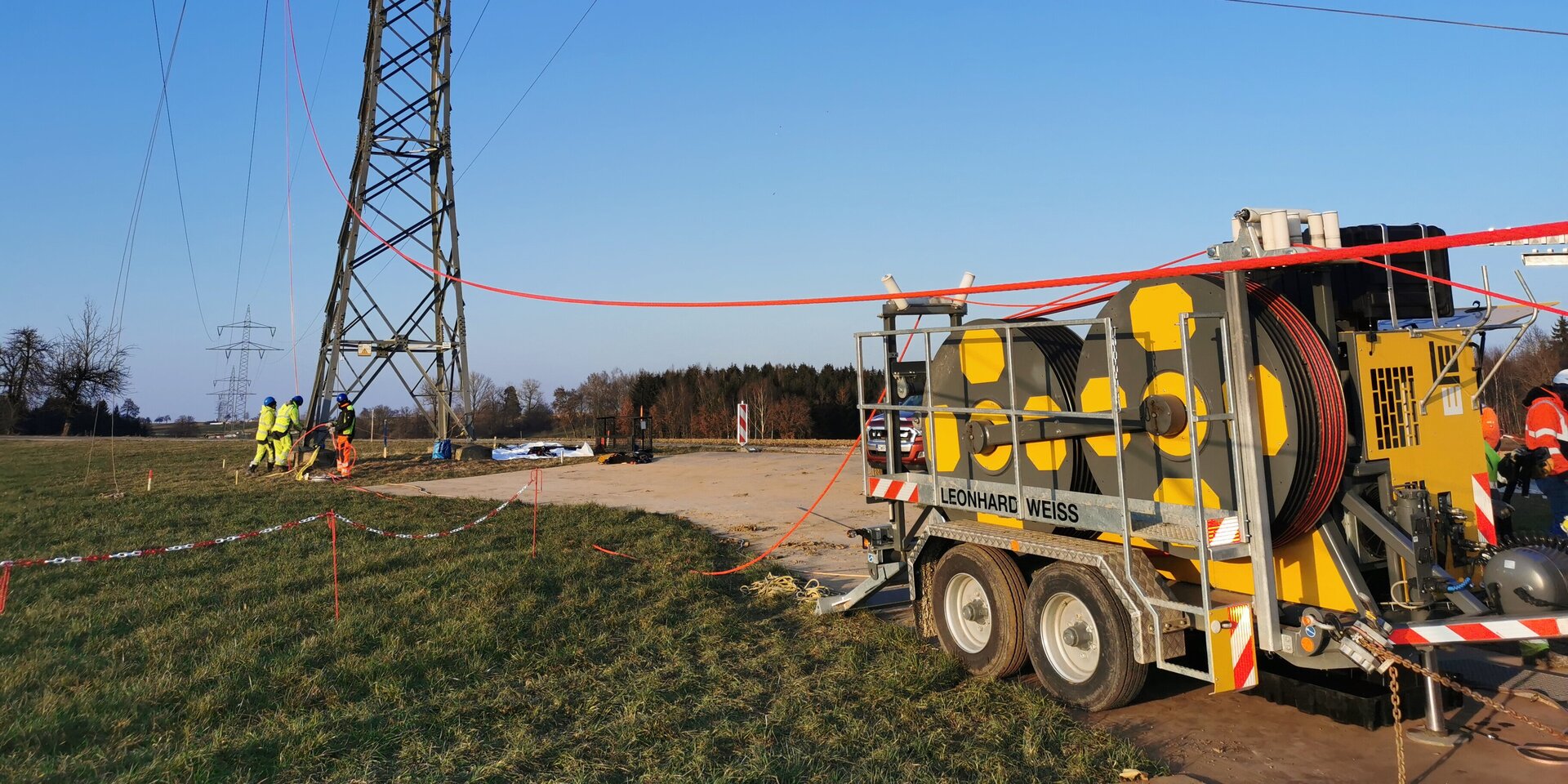 Die Szene zeigt Techniker, die an einem Strommast arbeiten, mit einem gelben Kabeltransportwagen und einem klaren, sonnigen Himmel im Hintergrund.