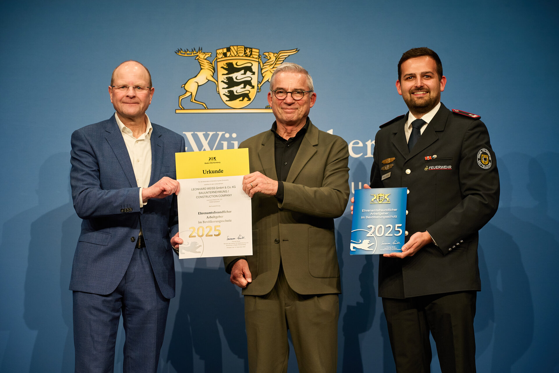Drei Männer in Anzügen halten Urkunden vor einer blauen Wand.