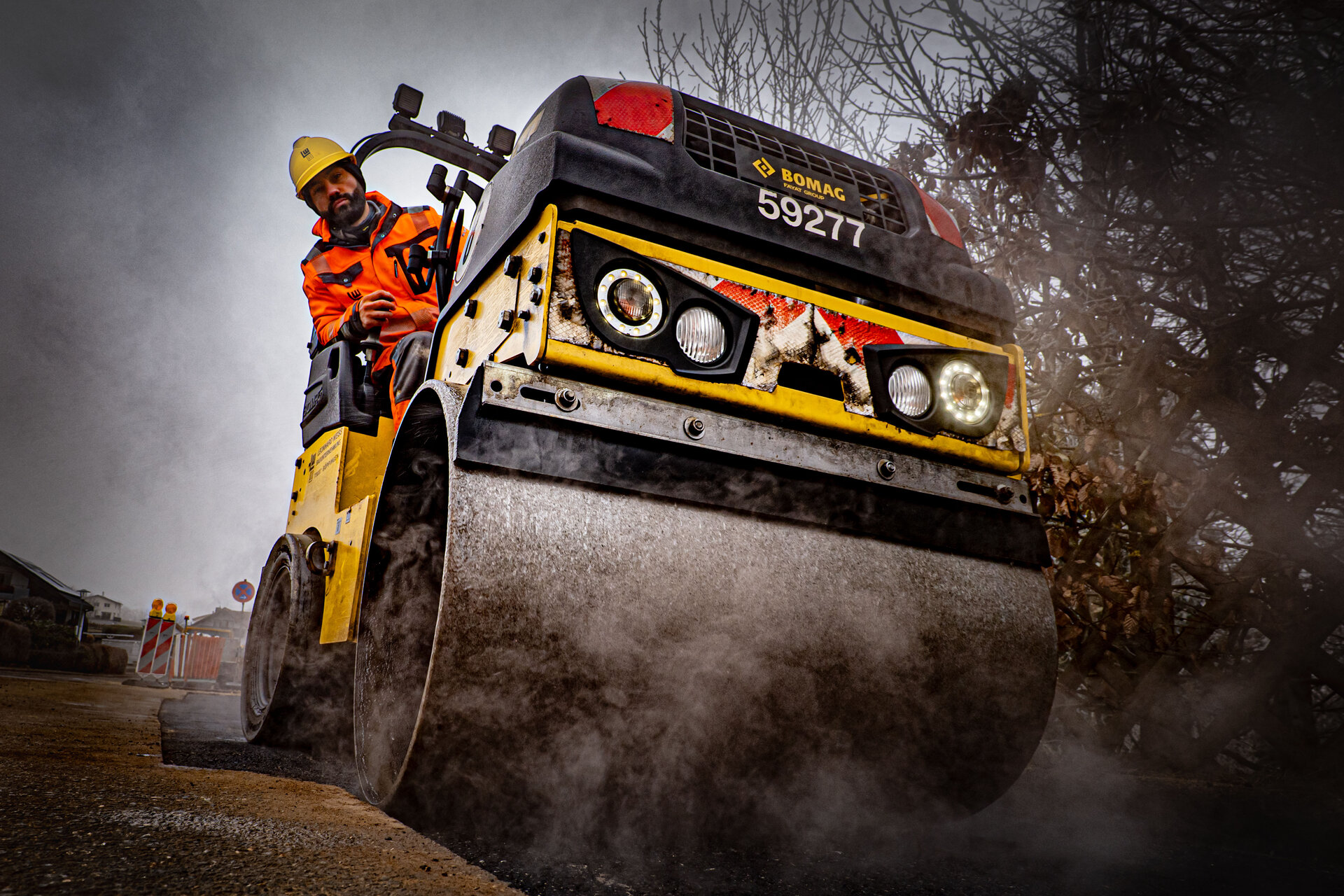 Ein Bauarbeiter in einem gelben Helm sitzt in einer Straßenwalze, die gerade Asphalt verdichtet, umgeben von einer mystischen Dunstschicht.