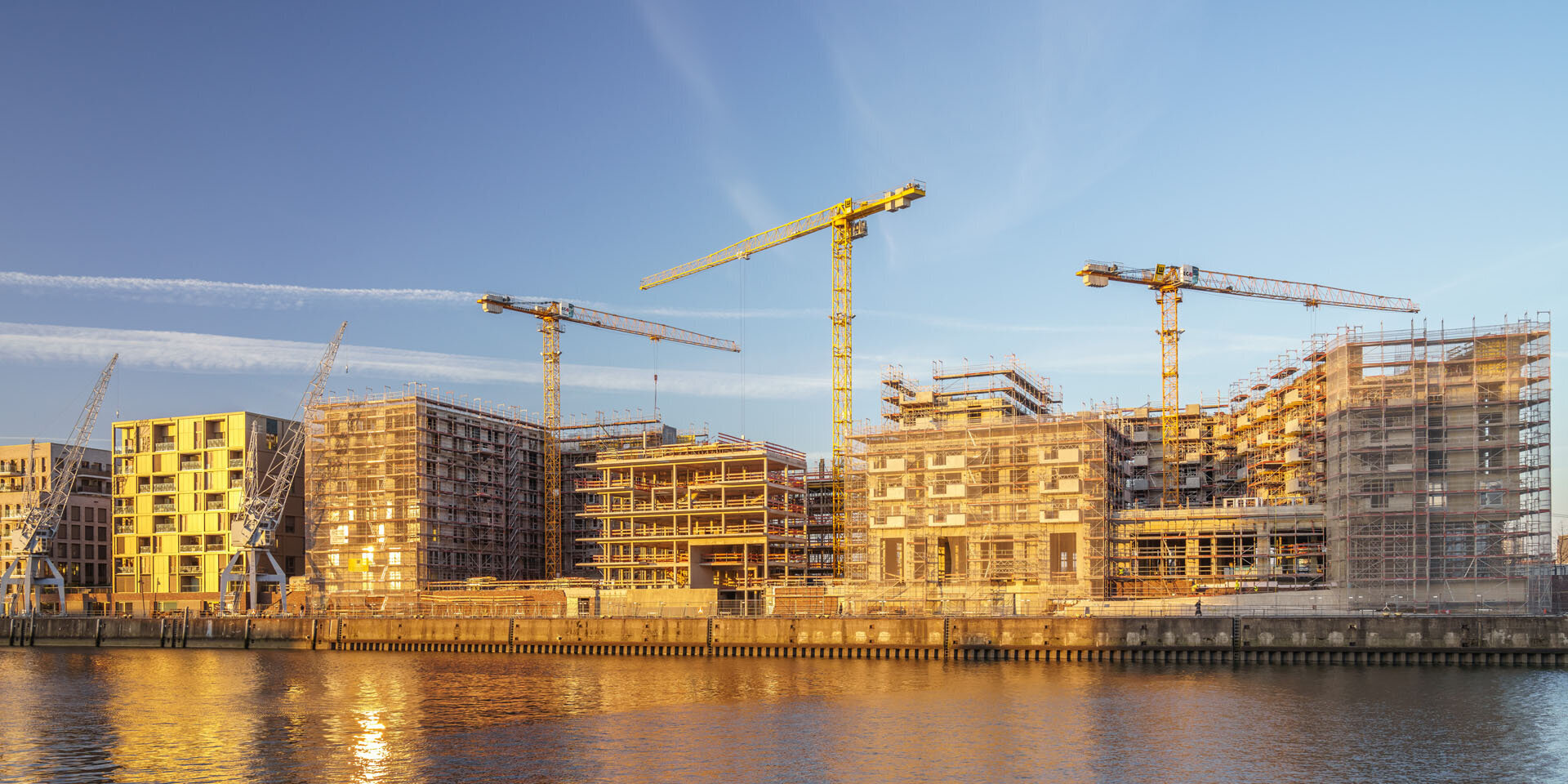 Eine Baustelle am Wasser mit mehreren Kränen und unvollendeten Gebäuden, die im warmen Licht der Abenddämmerung leuchten.