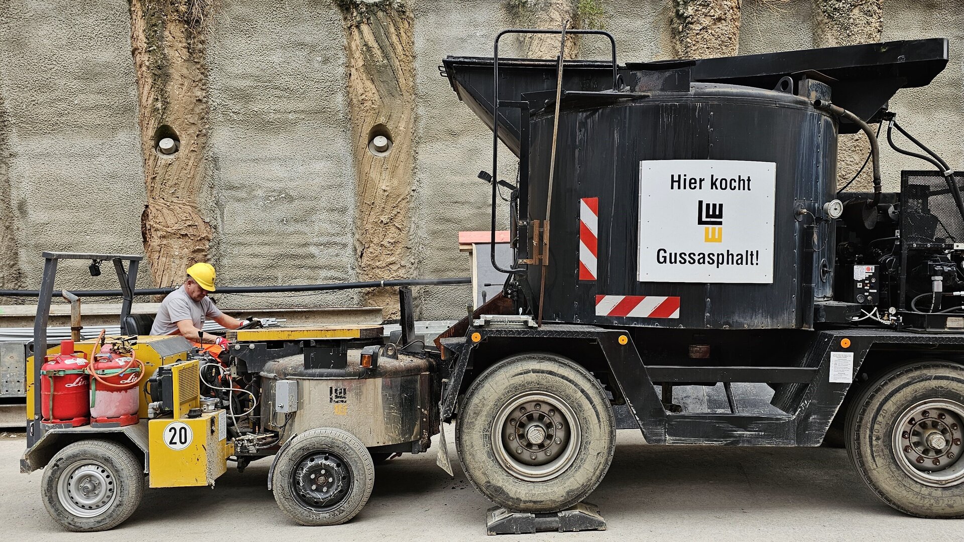 Ein Arbeiter in einem grauen T-Shirt und gelbem Helm bedient eine Maschine zur Asphaltproduktion auf einer Baustelle.