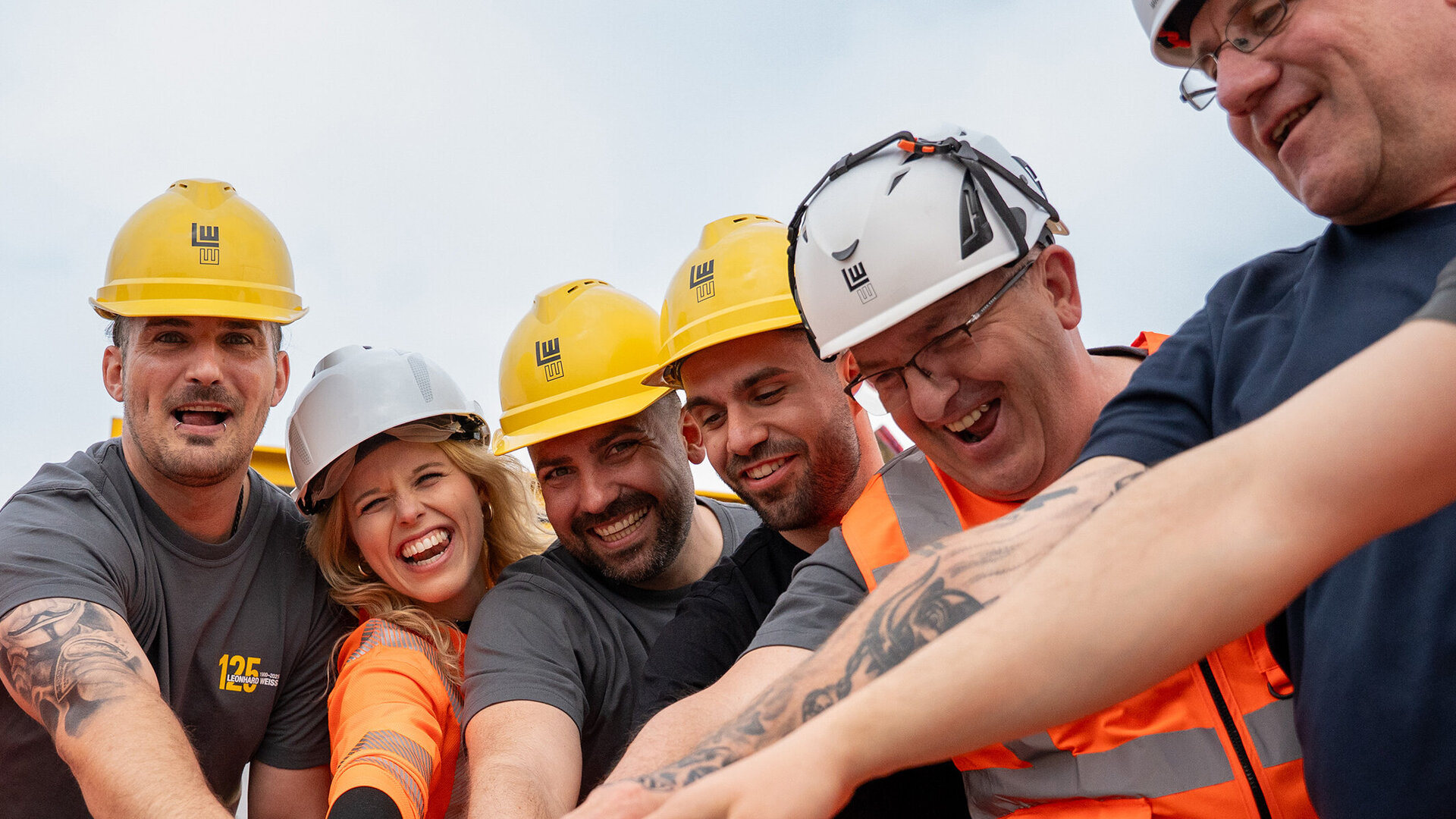 Die Mitarbeiter, gekleidet in orangefarbene Arbeitskleidung, lachen und zeigen ihre Verbundenheit durch das Zusammenlegen der Hände.