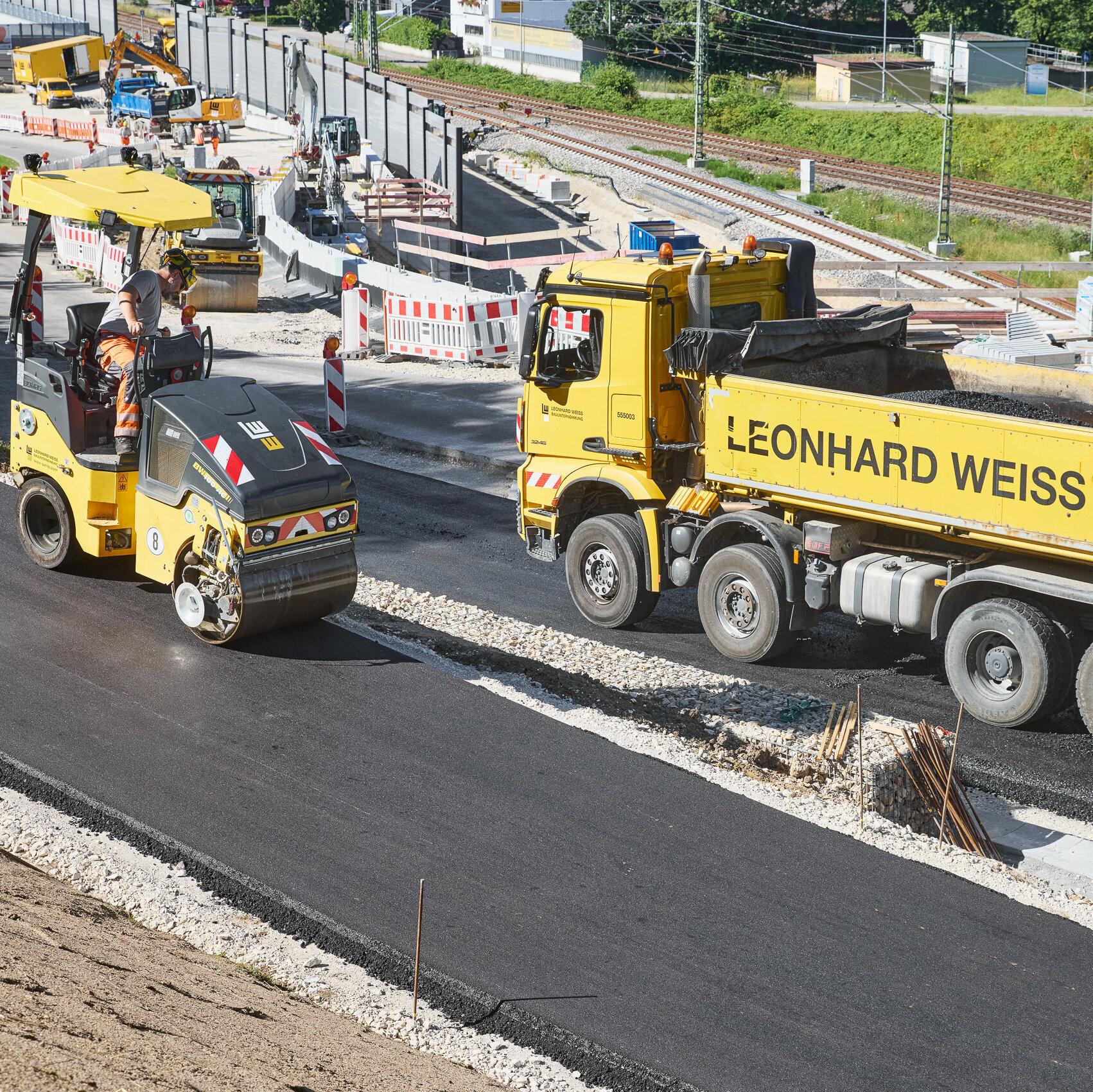 Ein Arbeiter steuert eine Walze über die frisch asphaltierte Straße, während im Hintergrund Bauarbeiten stattfinden.