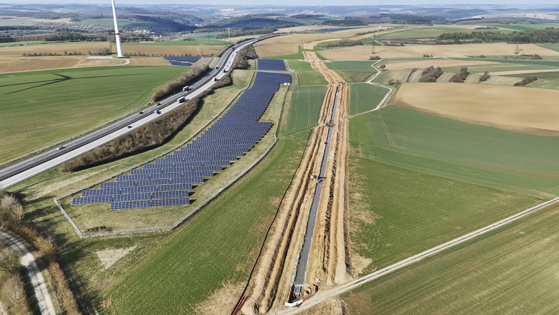 Eine Luftaufnahme von Bauarbeiten neben einer Autobahn, umgeben von grünen Feldern und Solarenergieanlagen.