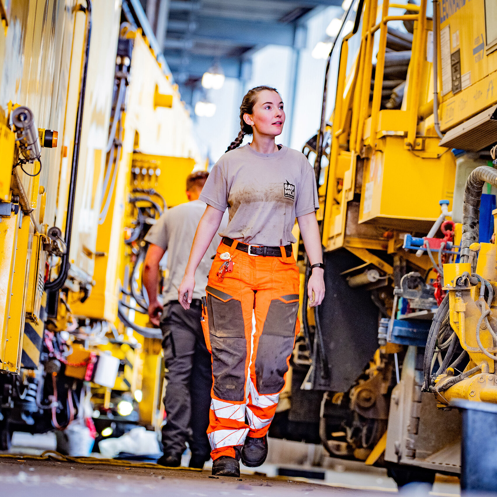 Die Arbeiterin mit Zöpfen trägt orangefarbene Hosen und schaut aufmerksam auf die Maschinen, während sie durch die Werkstatt geht.