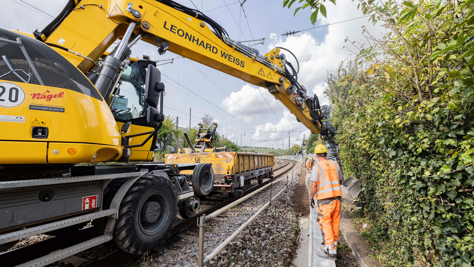 Ein gelber Schienenbagger arbeitet am Gleis-Kabelkanal, während ein Bauarbeiter in orangefarbener Kleidung die Arbeiten überwacht.