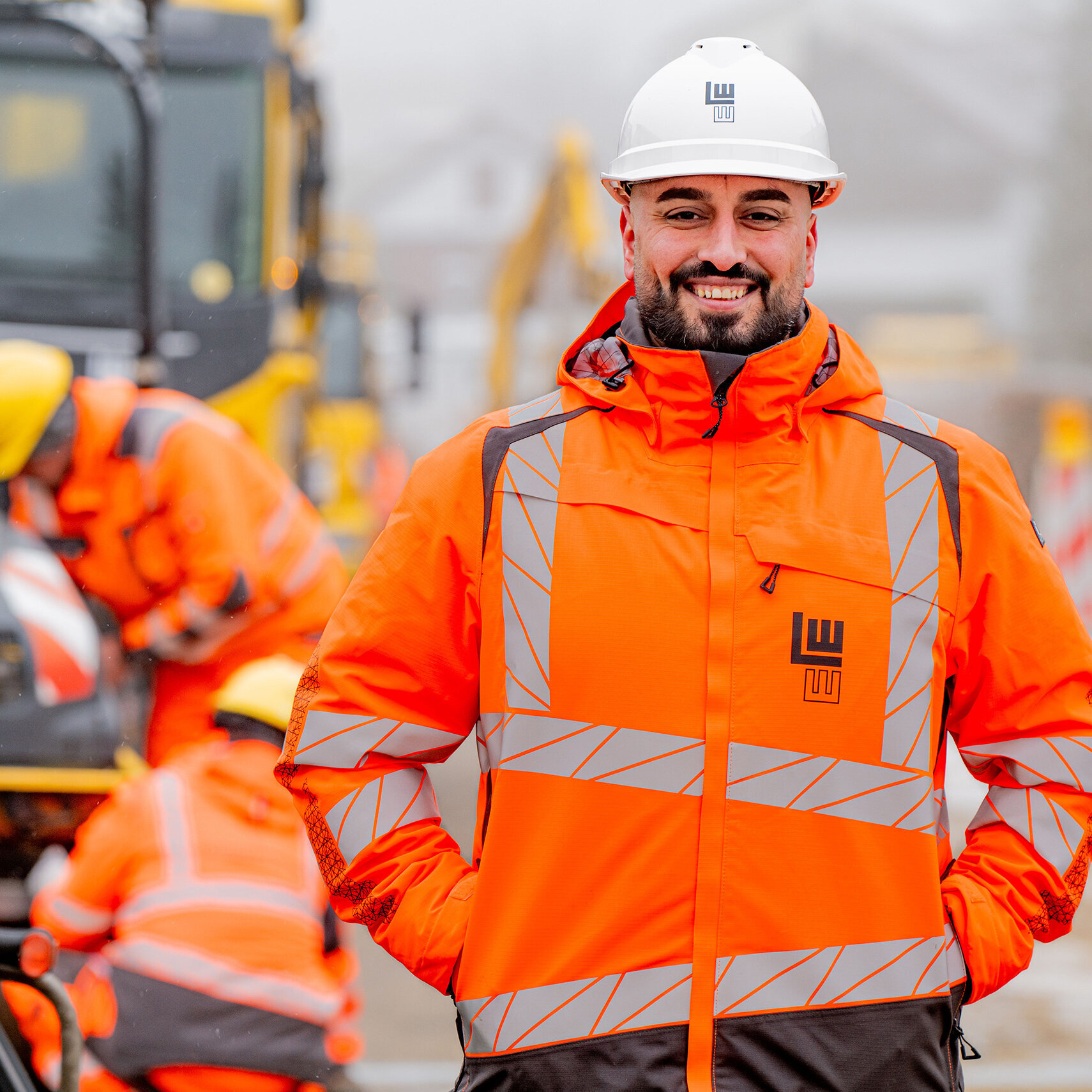 Ein lächelnder Bauarbeiter in leuchtend orangefarbener Arbeitskleidung steht vor einer Baustelle mit Maschinen und Kollegen im Hintergrund.
