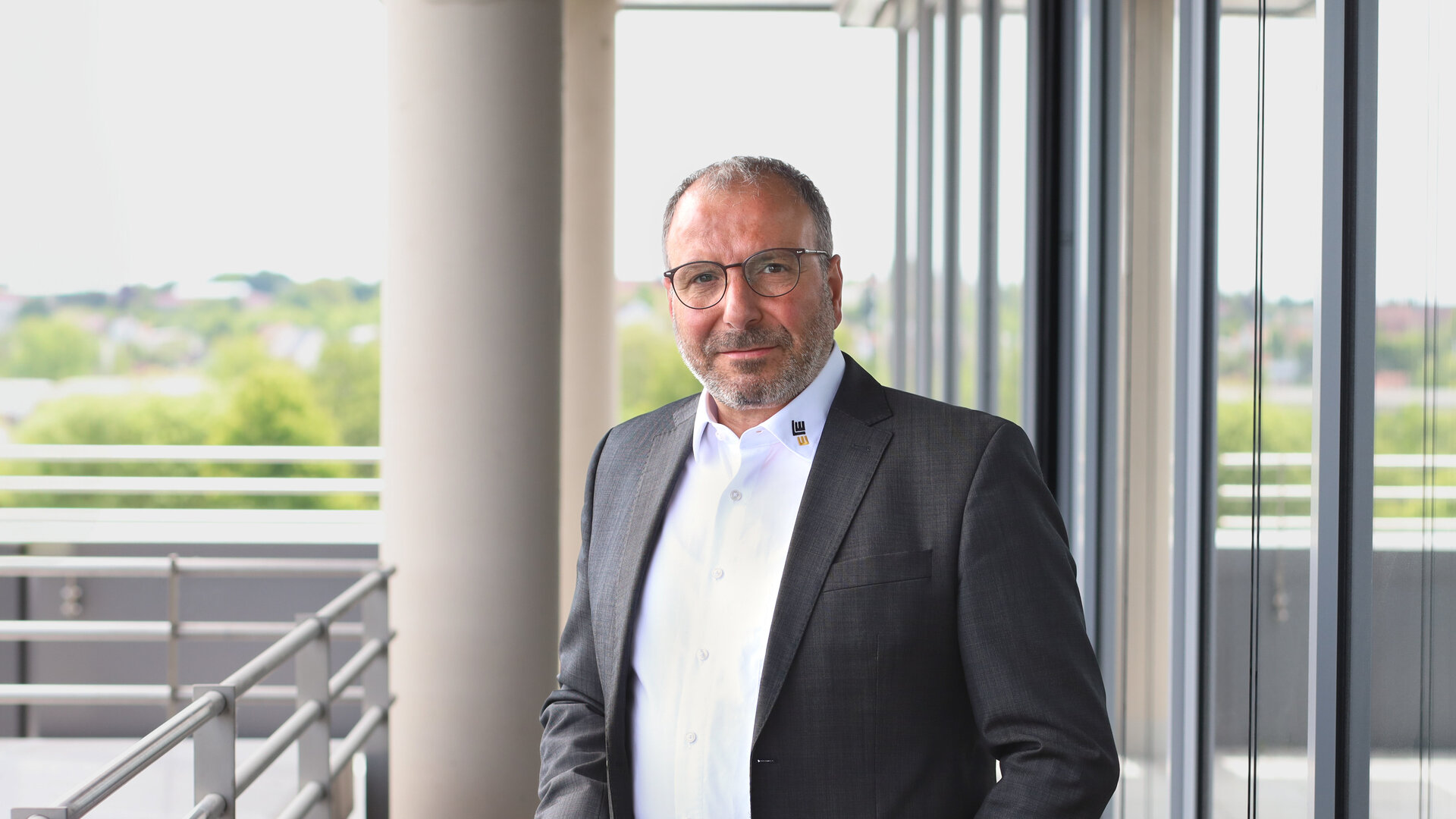 In einem stilvollen Bürogebäude posiert ein Mann in formeller Kleidung vor großen Fenstern mit natürlichem Licht.