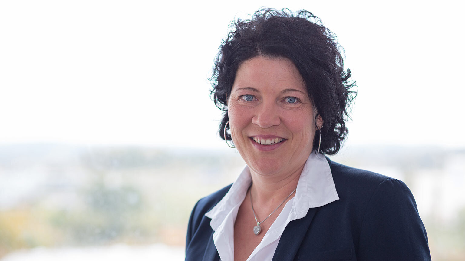 In einem hellen Raum steht eine Frau mit einem weißen Hemd und einem dunklen Blazer, die selbstbewusst in die Kamera lächelt.