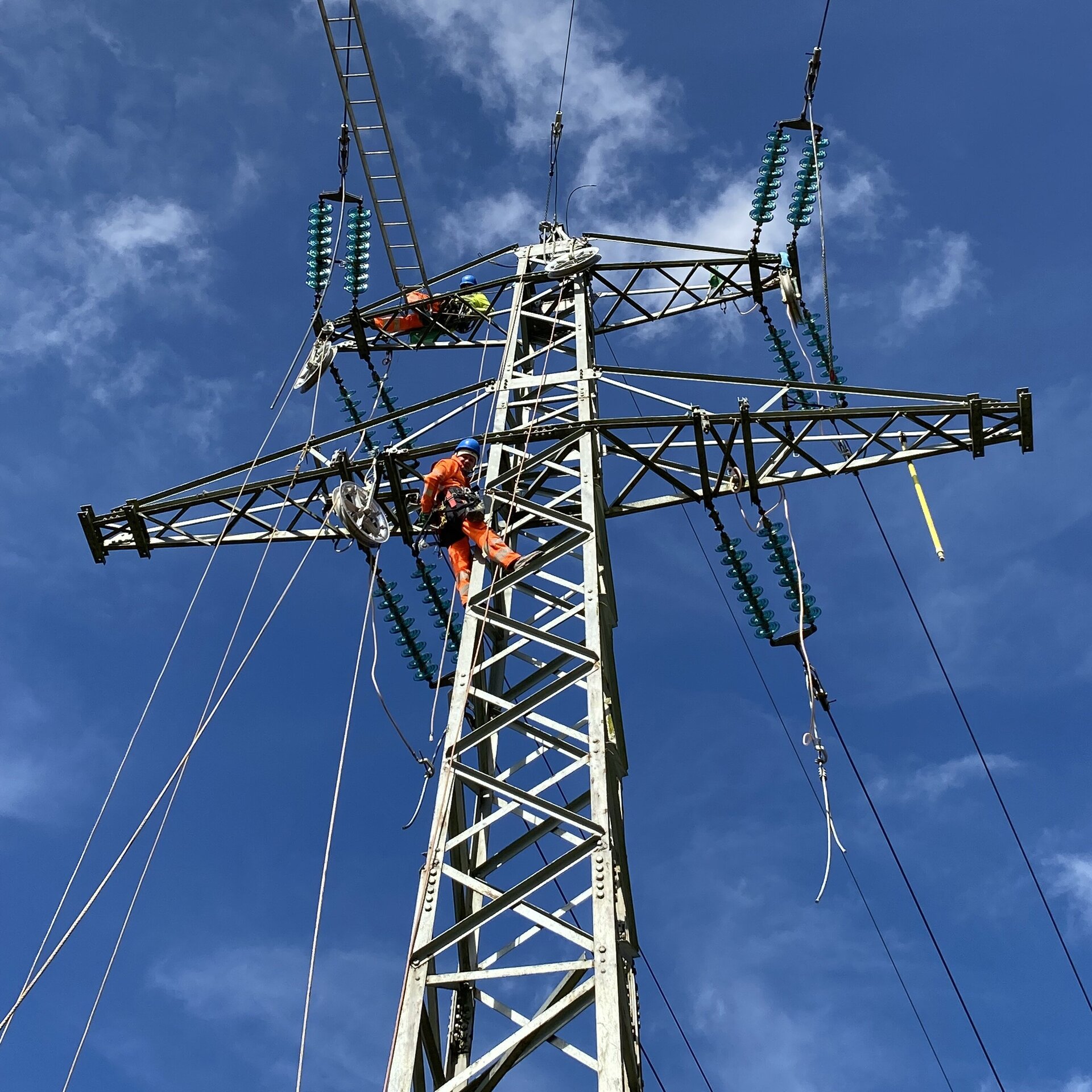Ein Arbeiter hängt an einem Strommast und repariert elektrische Leitungen, während die Wolken sanft über den Himmel ziehen.