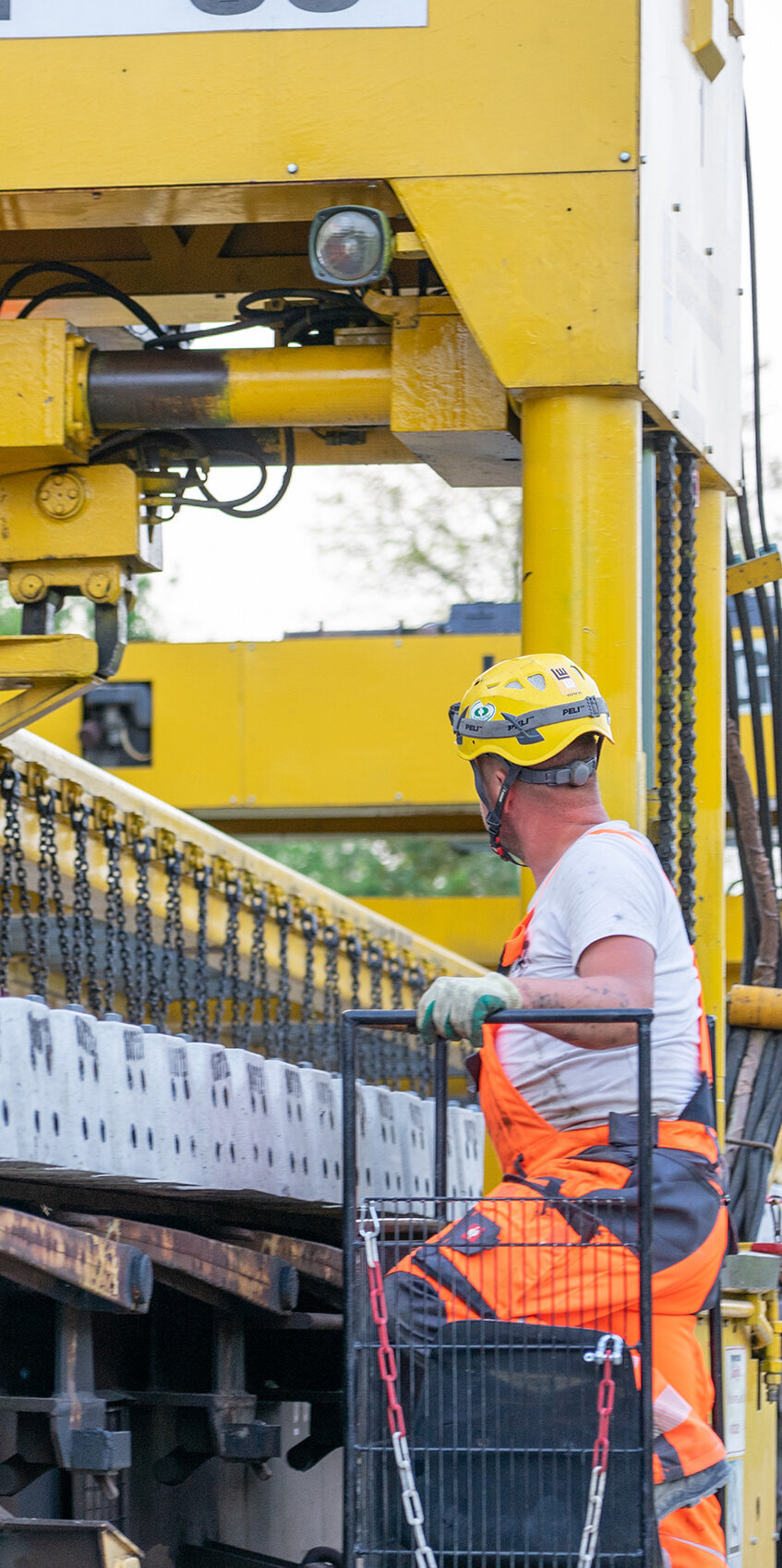 Ein Arbeiter in orangefarbener Arbeitskleidung überwacht den Einsatz eines Donelli – Gleisverlegeportalkrans, der Betonplatten hebt und bewegt.