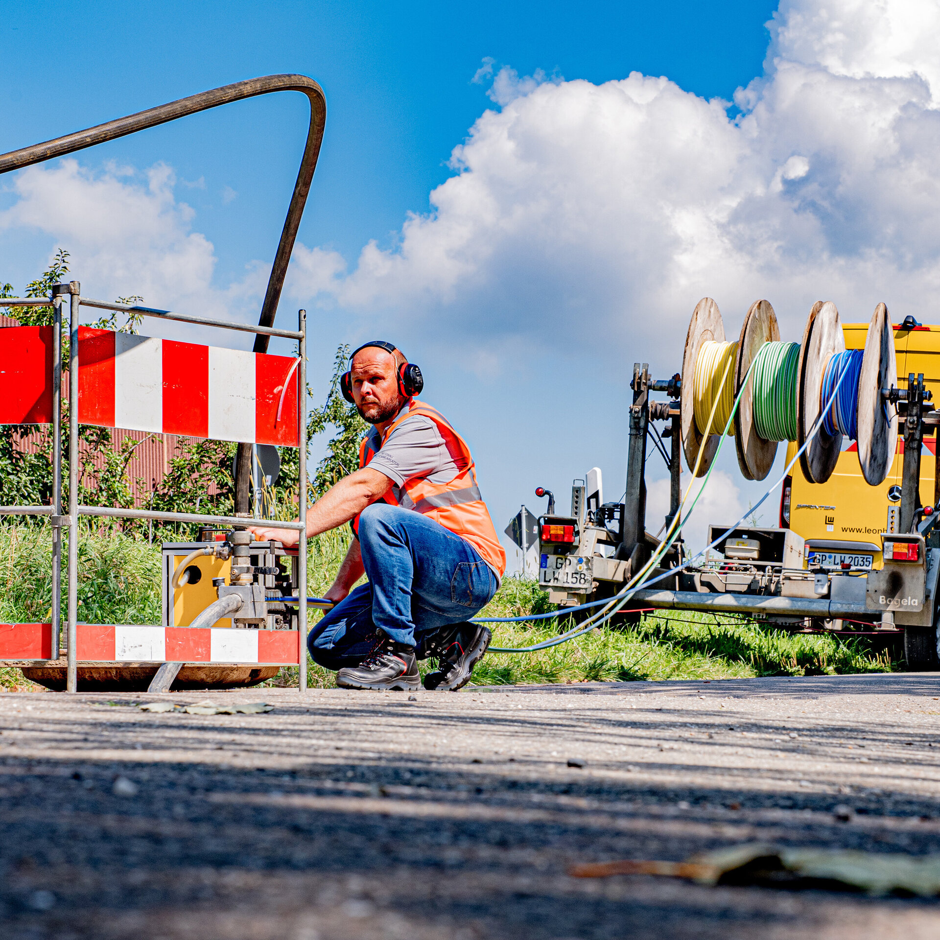 Bauarbeiter in Warnweste und Gehörschutz arbeitet an einer Straßenbaustelle. Neben ihm eine Absperrung und Kabeltechnik, im Hintergrund ein gelber Transporter mit großen Kabeltrommeln.