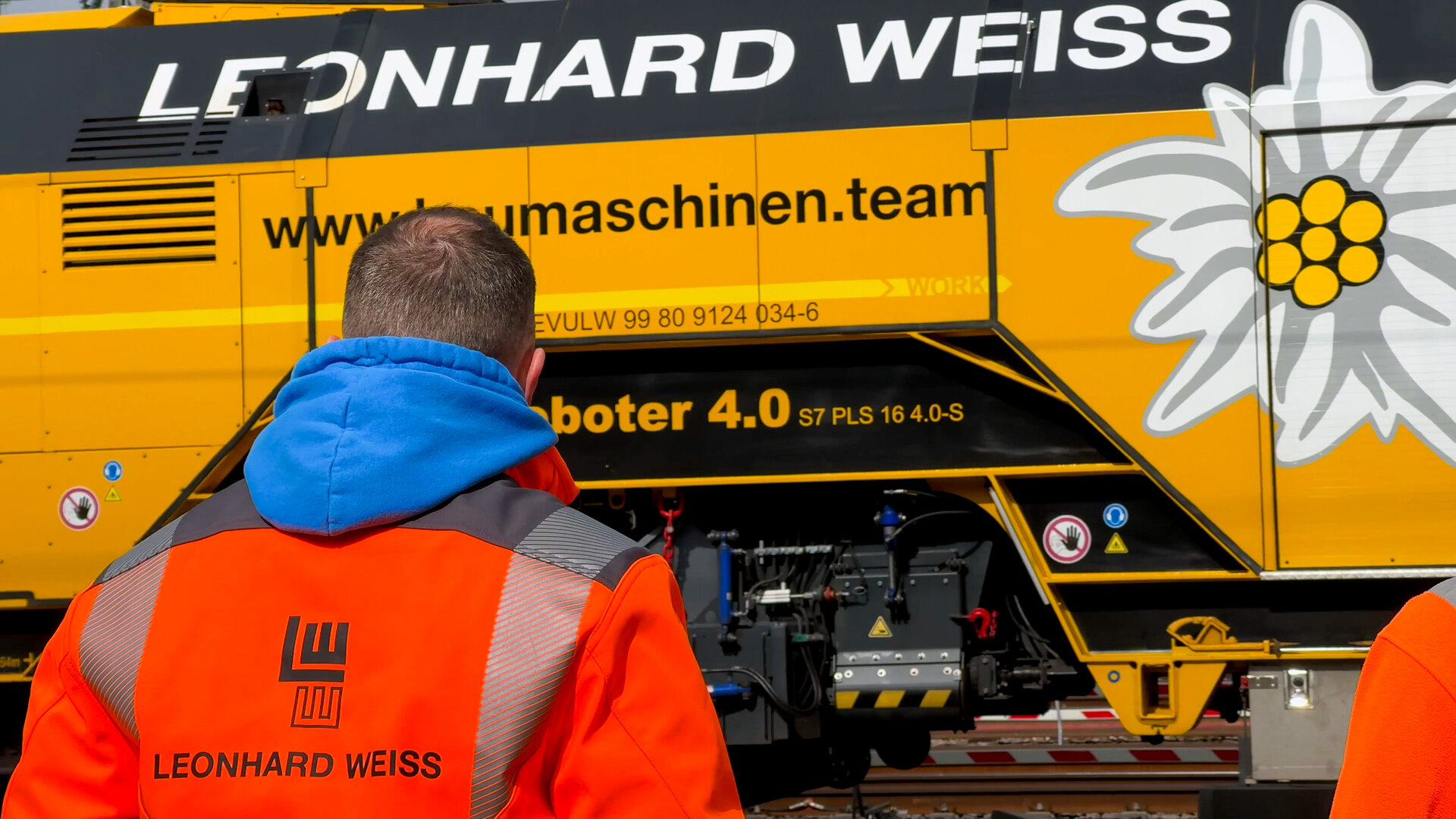 Ein Arbeiter in leuchtend orangefarbener Kleidung steht vor einer gelben Maschine mit dem Logo von Leonhard Weiss.