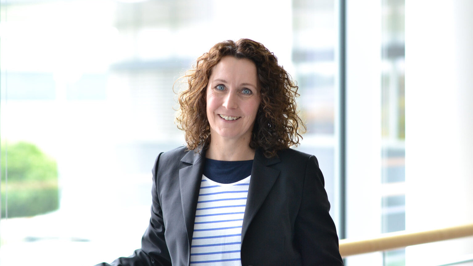 Porträt einer Frau mit lockigem braunem Haar, die lächelnd in die Kamera blickt. Sie trägt ein gestreiftes Shirt mit dunklem Blazer und steht in einem hellen, modernen Gebäude.