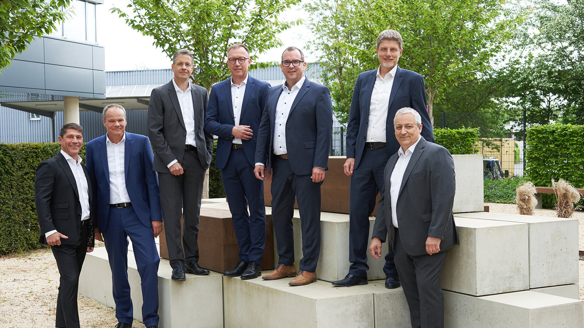 Group photo of the management team in suits standing on an outdoor concrete structure, surrounded by trees and a modern building.
