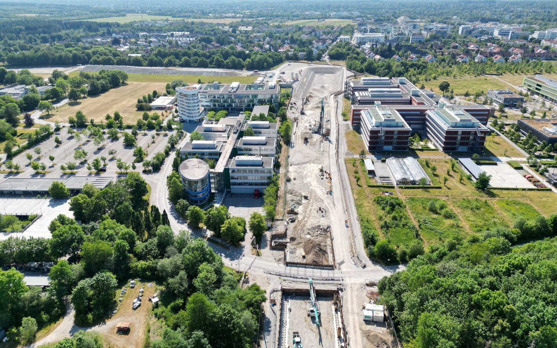 Im Bild sind moderne Bürogebäude und eine Baustelle zu sehen, umgeben von viel Grün und einer Waldlandschaft im Hintergrund.