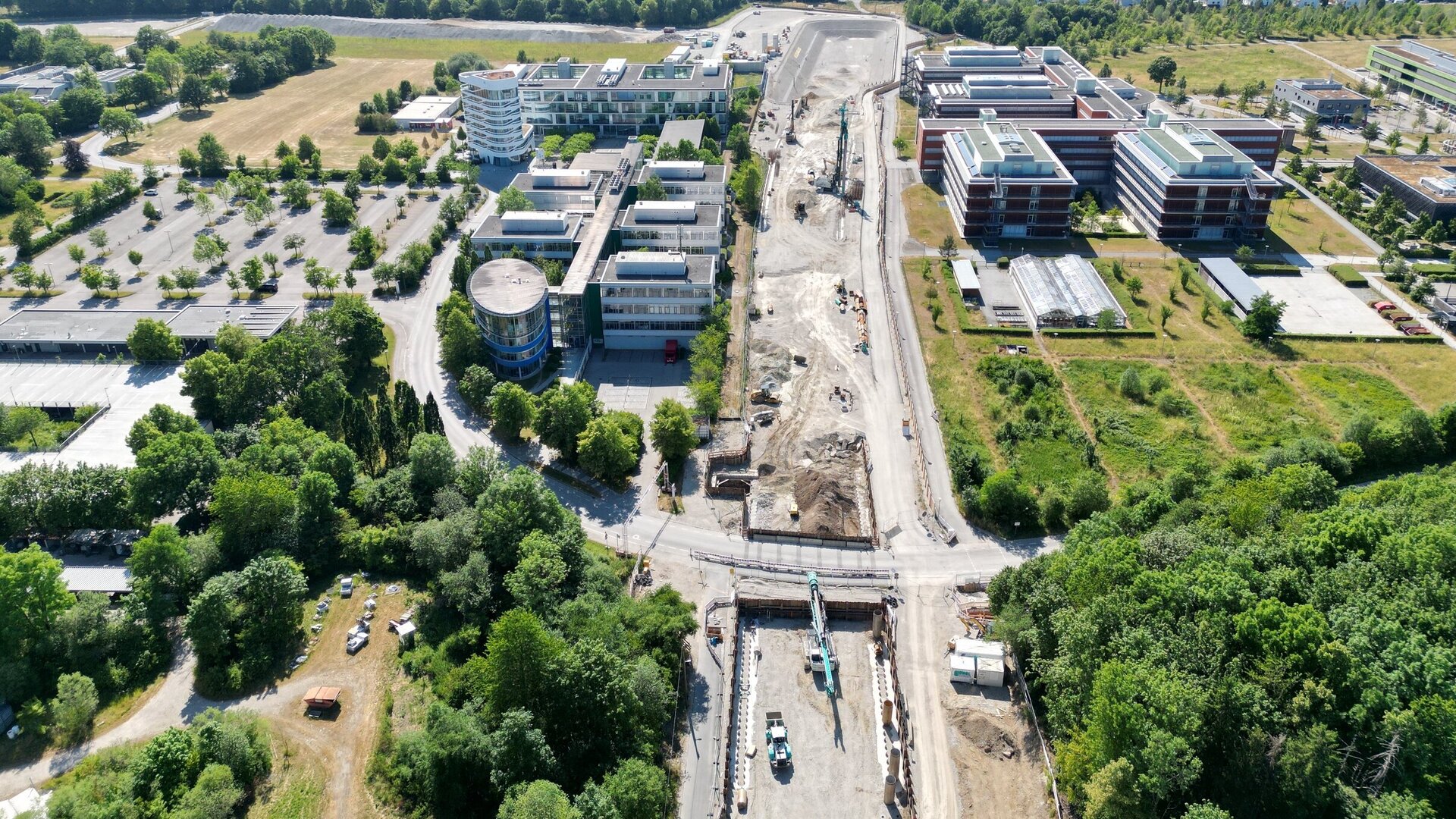 Im Bild sind moderne Bürogebäude und eine Baustelle zu sehen, umgeben von viel Grün und einer Waldlandschaft im Hintergrund.