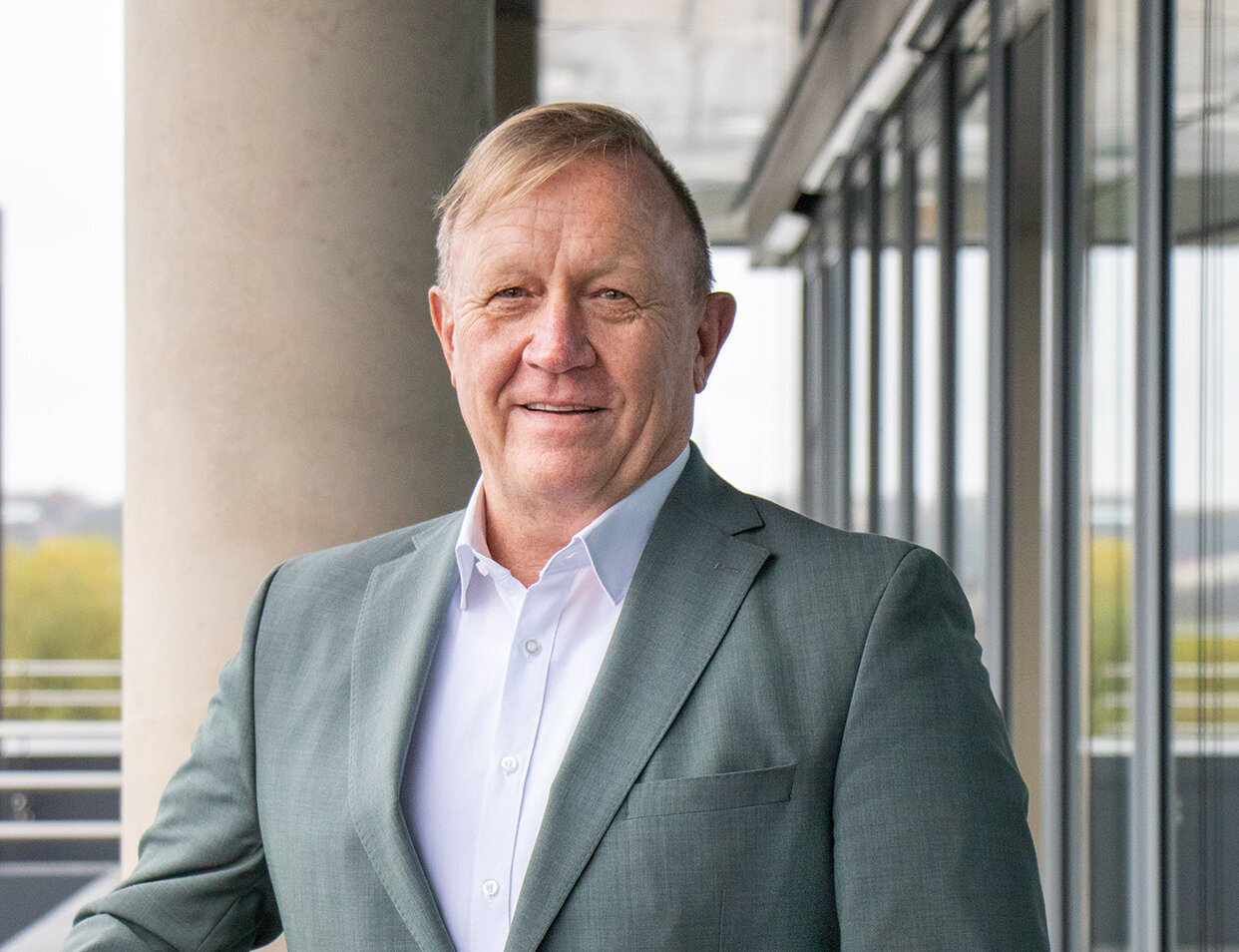 Vor einer modernen Glasfassade steht ein Mann, der in einem grauen Blazer und einem weißen Hemd gekleidet ist.