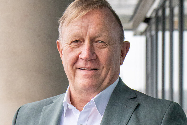 Vor einer modernen Glasfassade steht ein Mann, der in einem grauen Blazer und einem weißen Hemd gekleidet ist.