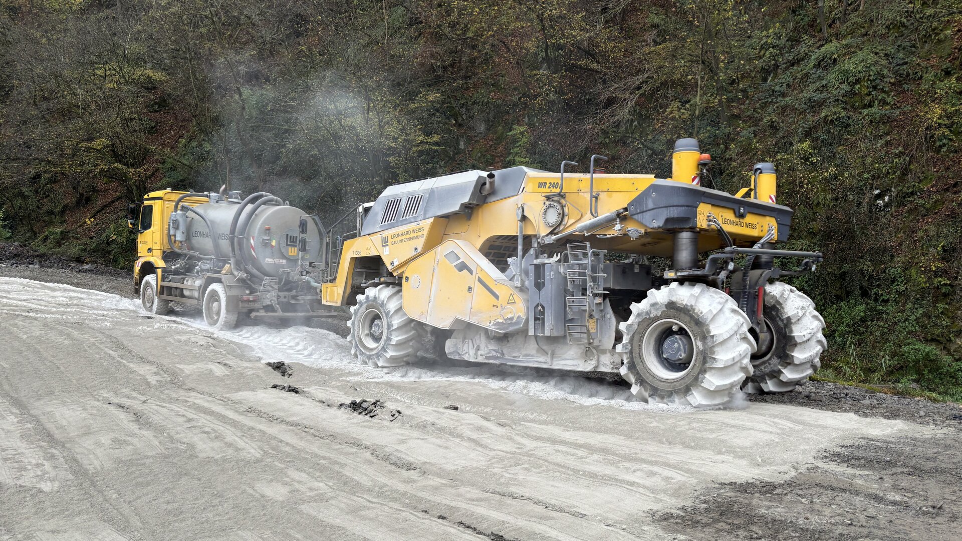 Ein gelbes Straßenbaugerät und ein Lkw sind auf einer Baustelle aktiv, um den Untergrund für eine neue Straße vorzubereiten und zu stabilisieren.