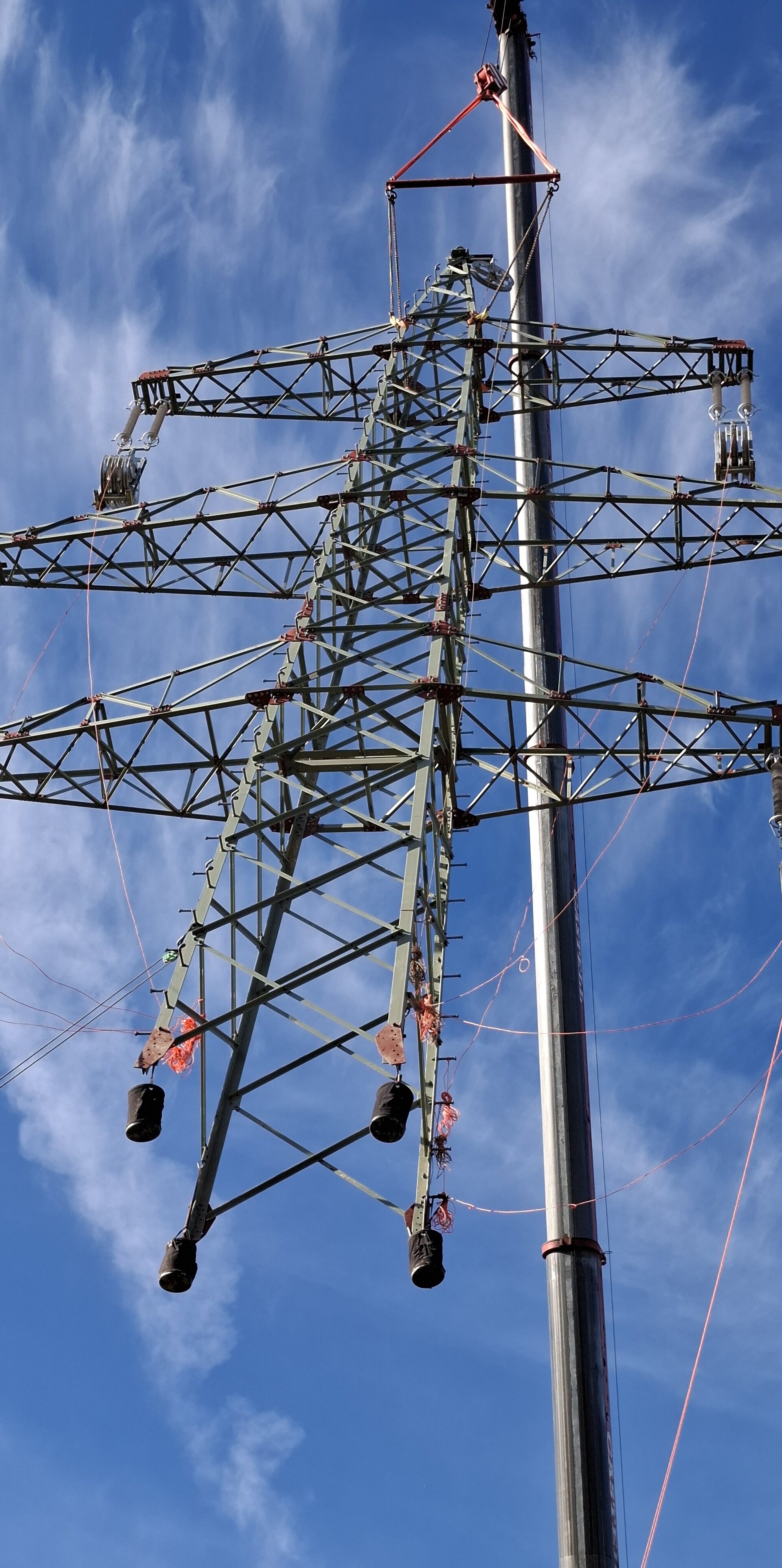 Ein Hochspannungsmast wird mit Hilfe eines Krans angehoben und montiert. Der Stahlgittermast ist mit Seilen und Hebegurten gesichert und schwebt teilweise in der Luft vor blauem Himmel.