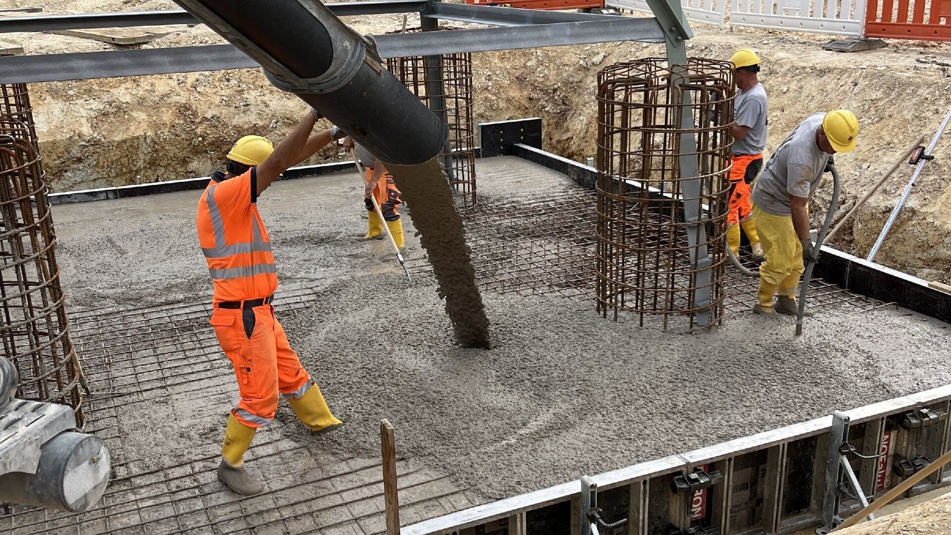 Die Szene zeigt Bauarbeiter, die Beton in eine vorbereitete Schalung gießen, um das Fundament für einen Strommasten zu erstellen.