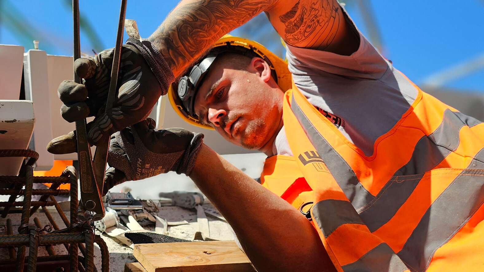 Ein Bauarbeiter in orangefarbener Sicherheitskleidung bindet im Freien mit einer Zange einen Bewehrungskorb.